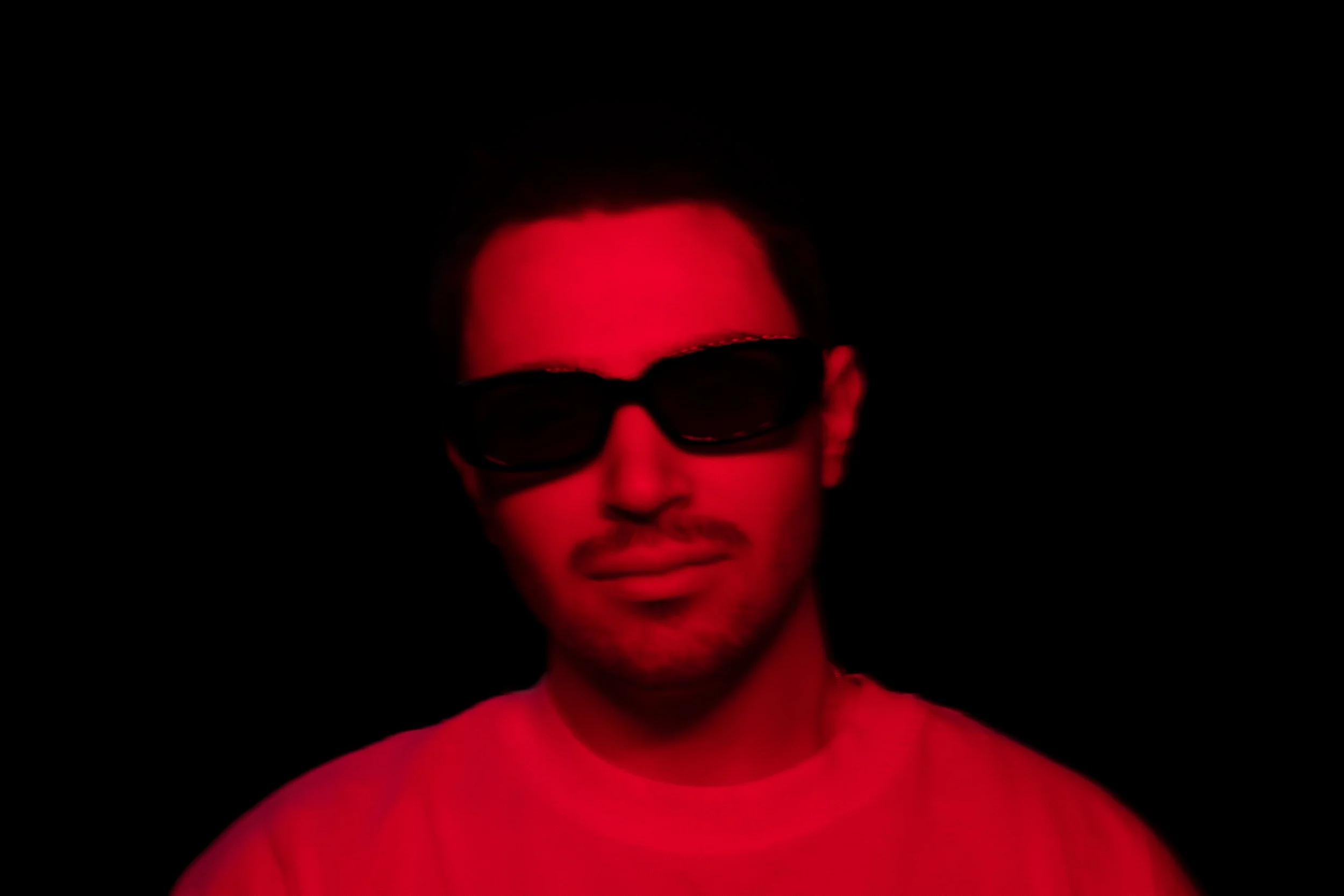A person with short dark hair wearing sunglasses and a pink shirt, illuminated by red light against a black background.