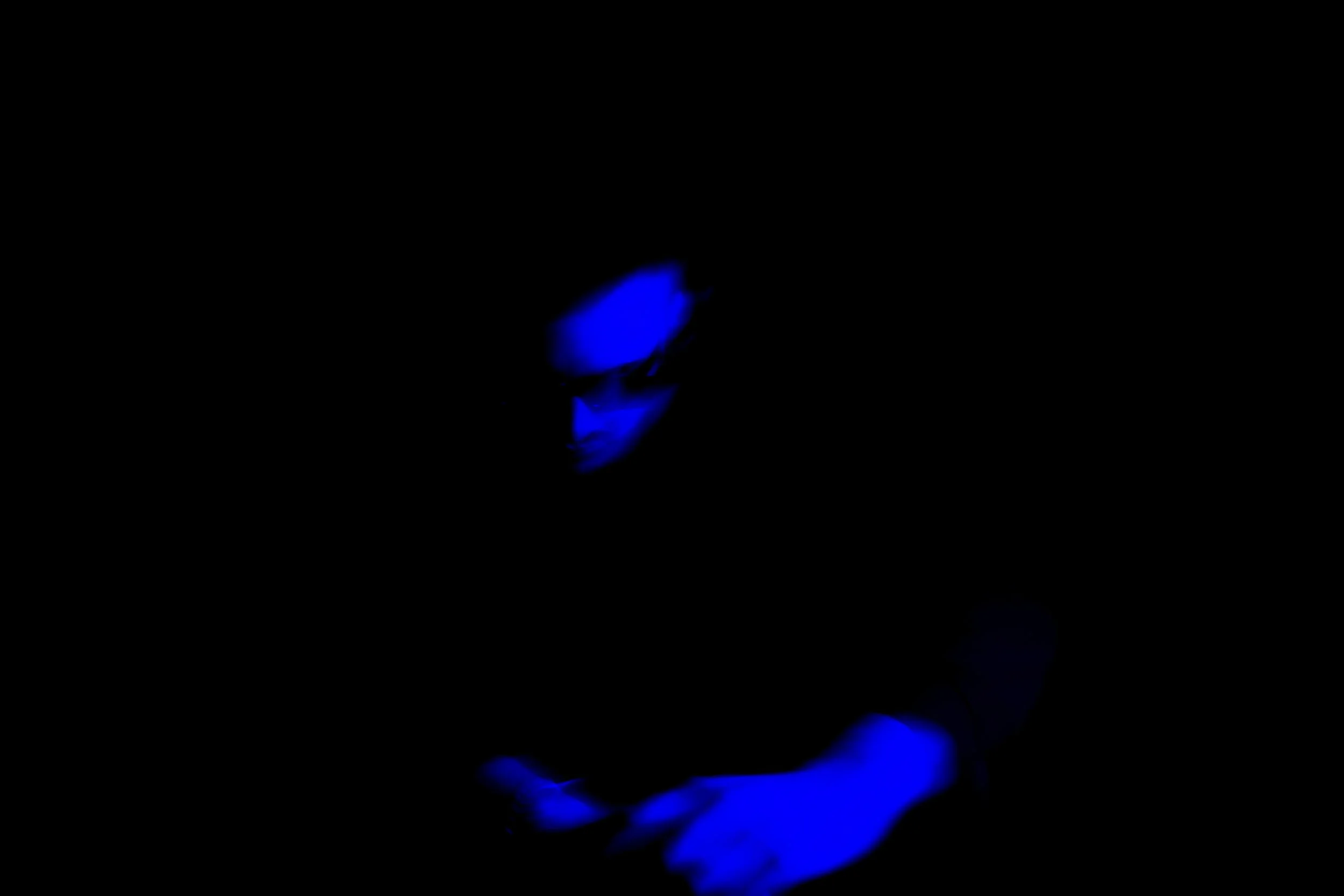 Person with glasses and a hat looking at a phone, illuminated by blue light in darkness.