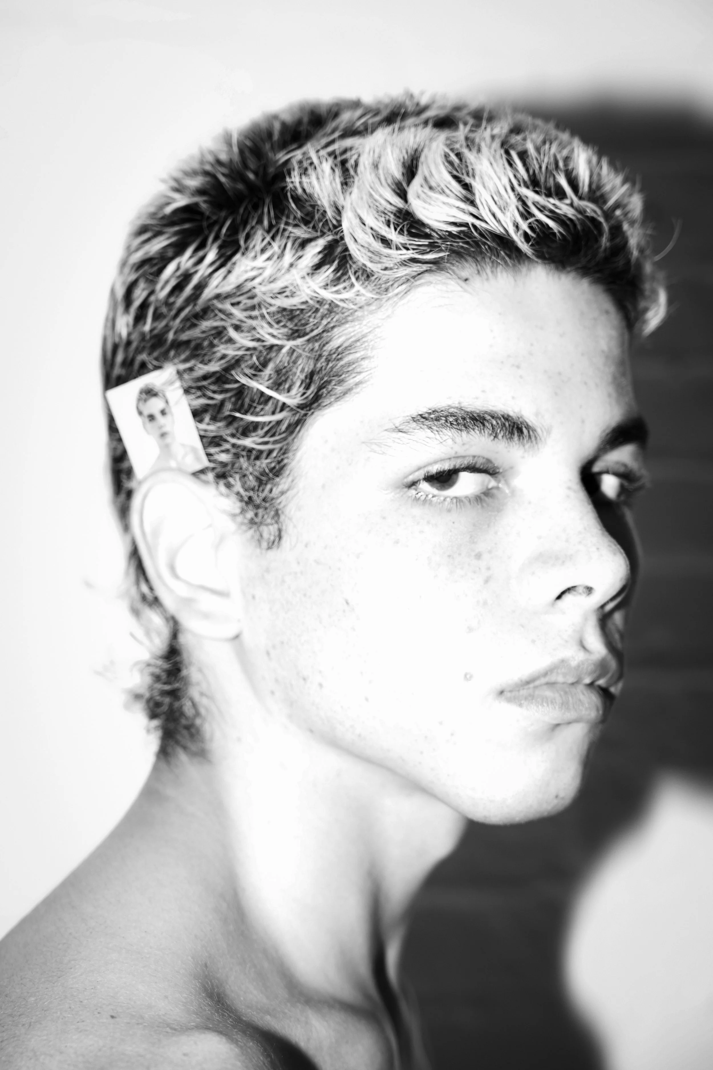 Black and white portrait of a young woman with short, wavy hair and freckles, with a small picture of her own face tucked behind her ear.