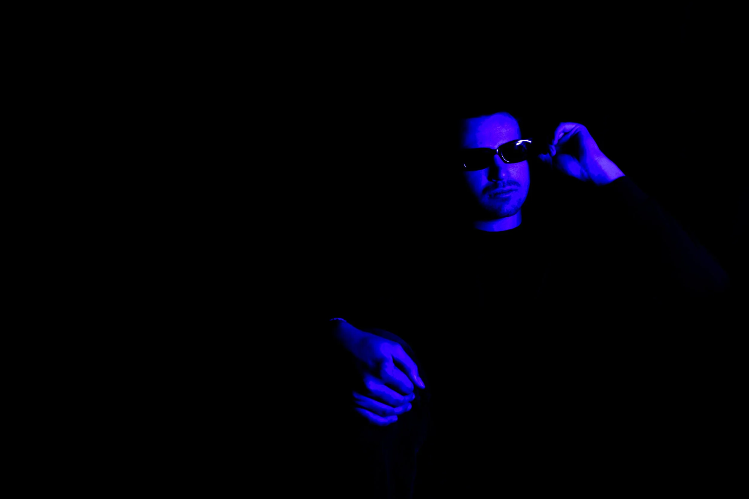 A man wearing dark sunglasses and a black shirt, illuminated by blue light, adjusting his sunglasses against a dark background.