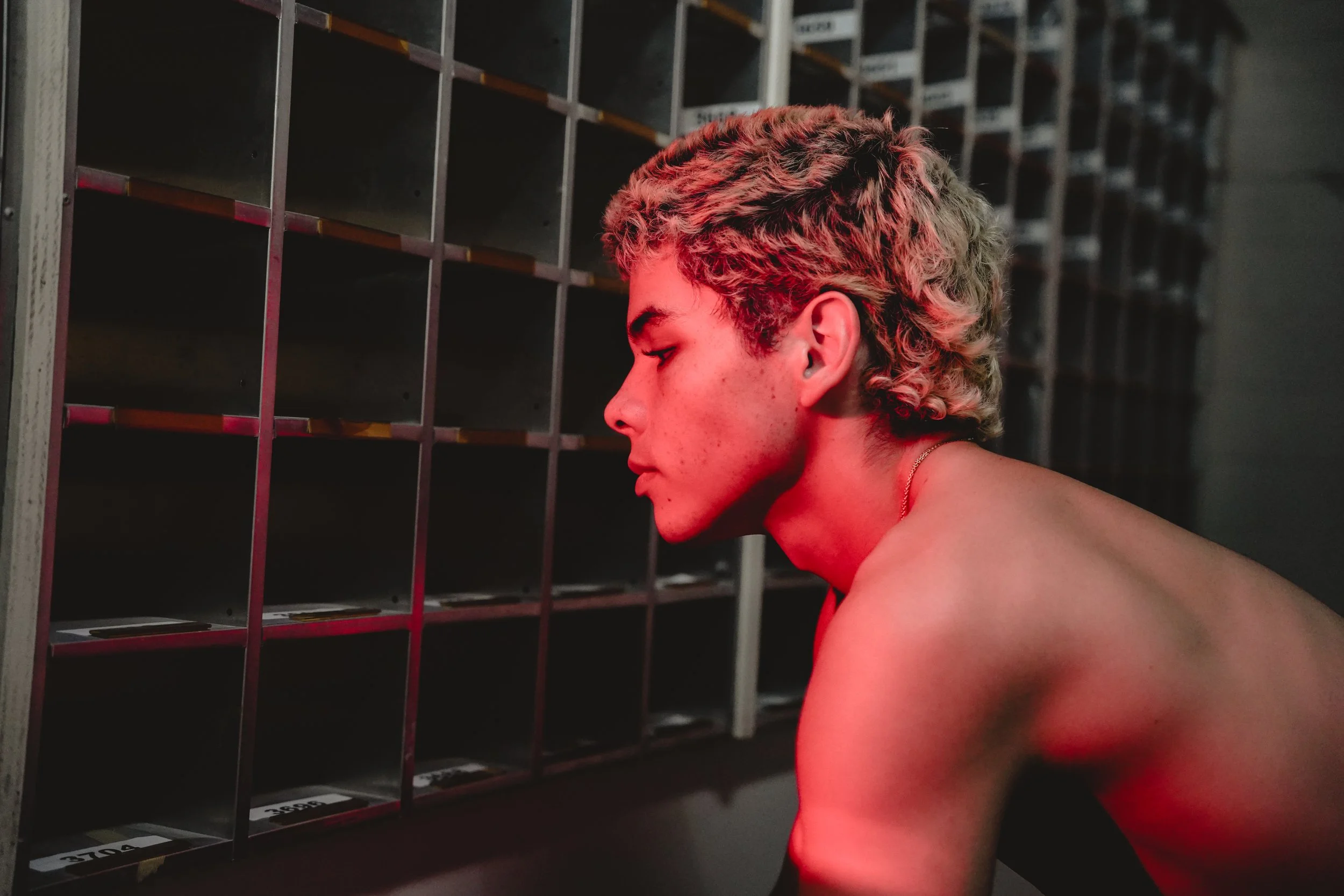 A young person with curly blonde hair and no shirt, looking at empty mailboxes or lockers in a dimly lit space with a red hue.