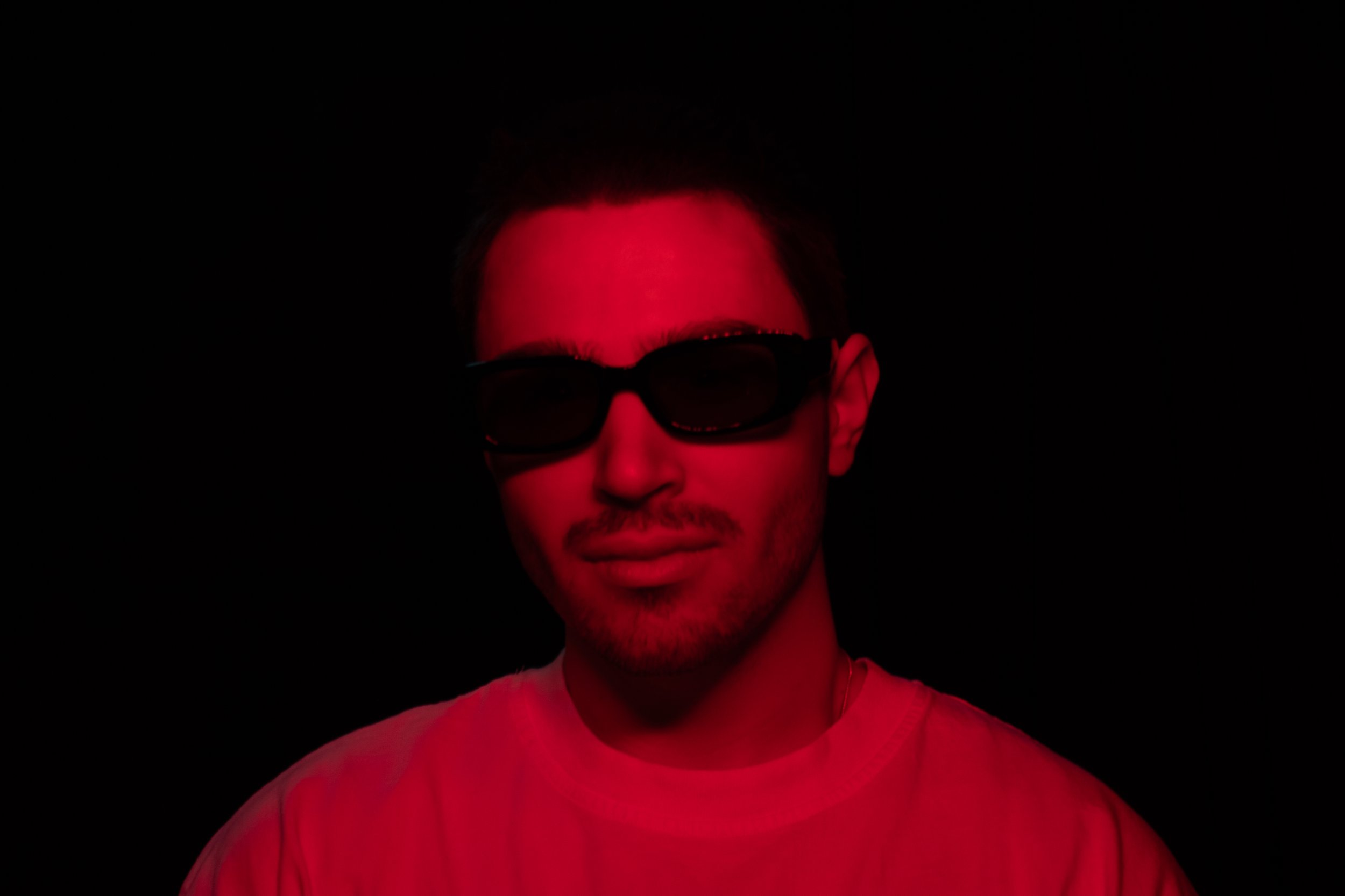 A man wearing sunglasses and a white shirt is illuminated by red lighting against a black background.