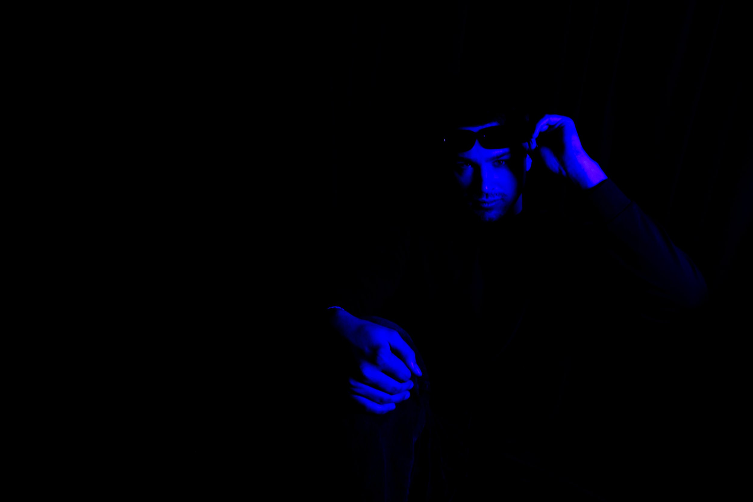 A person wearing sunglasses with blue lighting, adjusting glasses with one hand and resting the other hand on their knee in a dark environment.