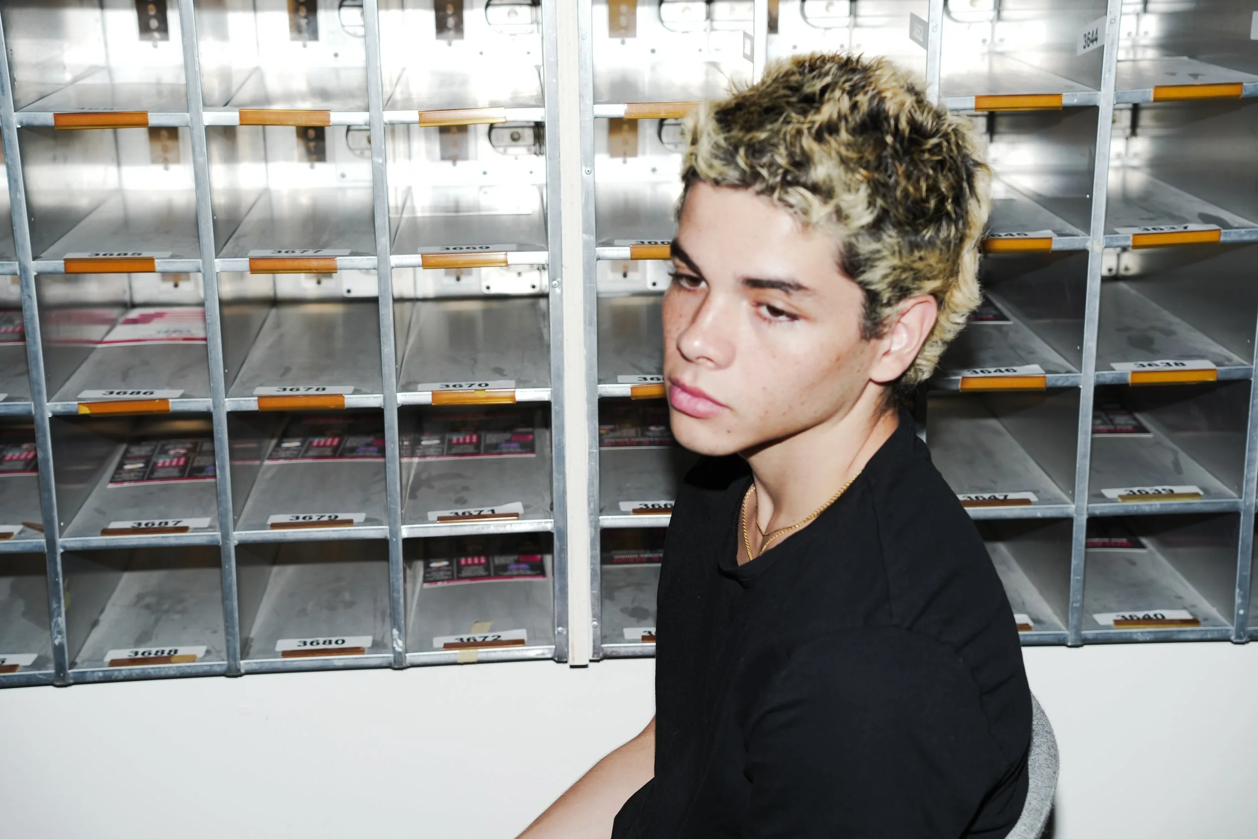 A young man with blonde, curly hair and a black shirt sitting in front of a metal storage locker with empty compartments and plastic trays.