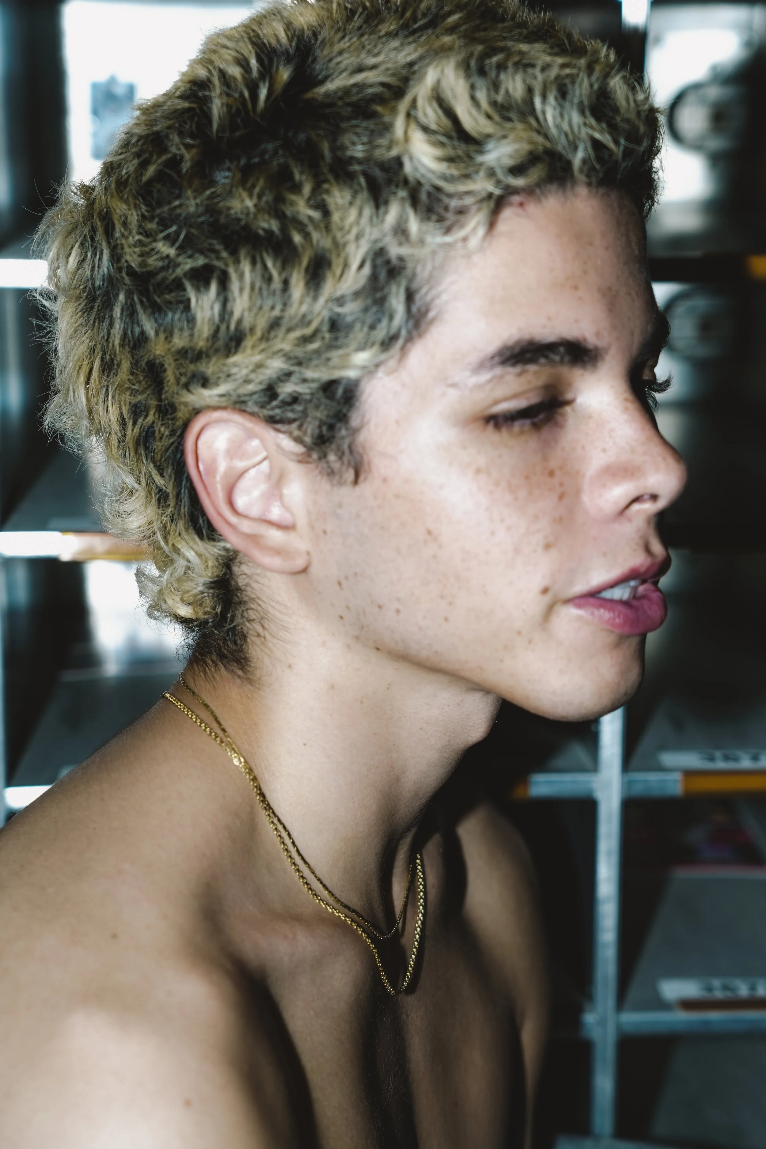 Close-up side profile of a young person with short, curly, blonde hair, freckles, wearing two gold necklaces, with a background of metal shelves and magazines.