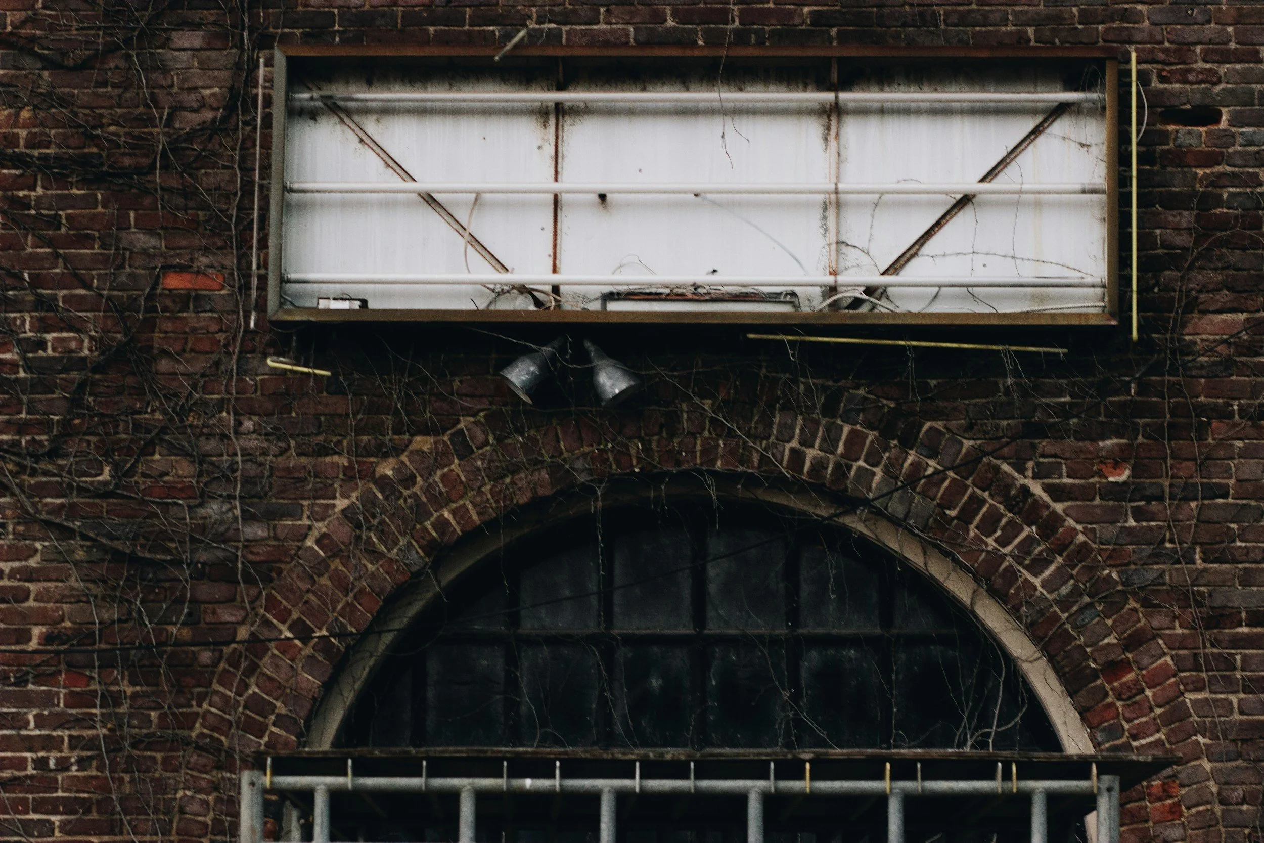A brick building with an arched window, some vines, and a metal security grille in front. There is an old, white, rusted sign or billboard above the window, with two gray spotlights below it.