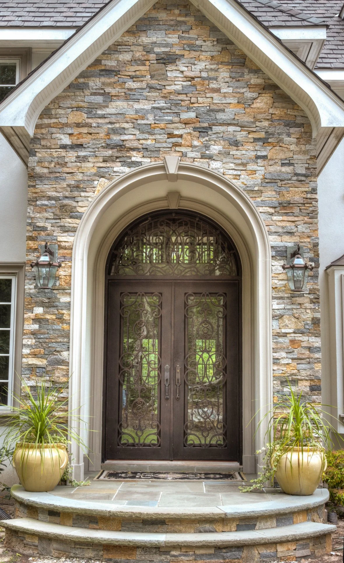 Front entrance of a house with double black ornate metal doors, stone facade, and two gold planters with green plants on each side of the steps.