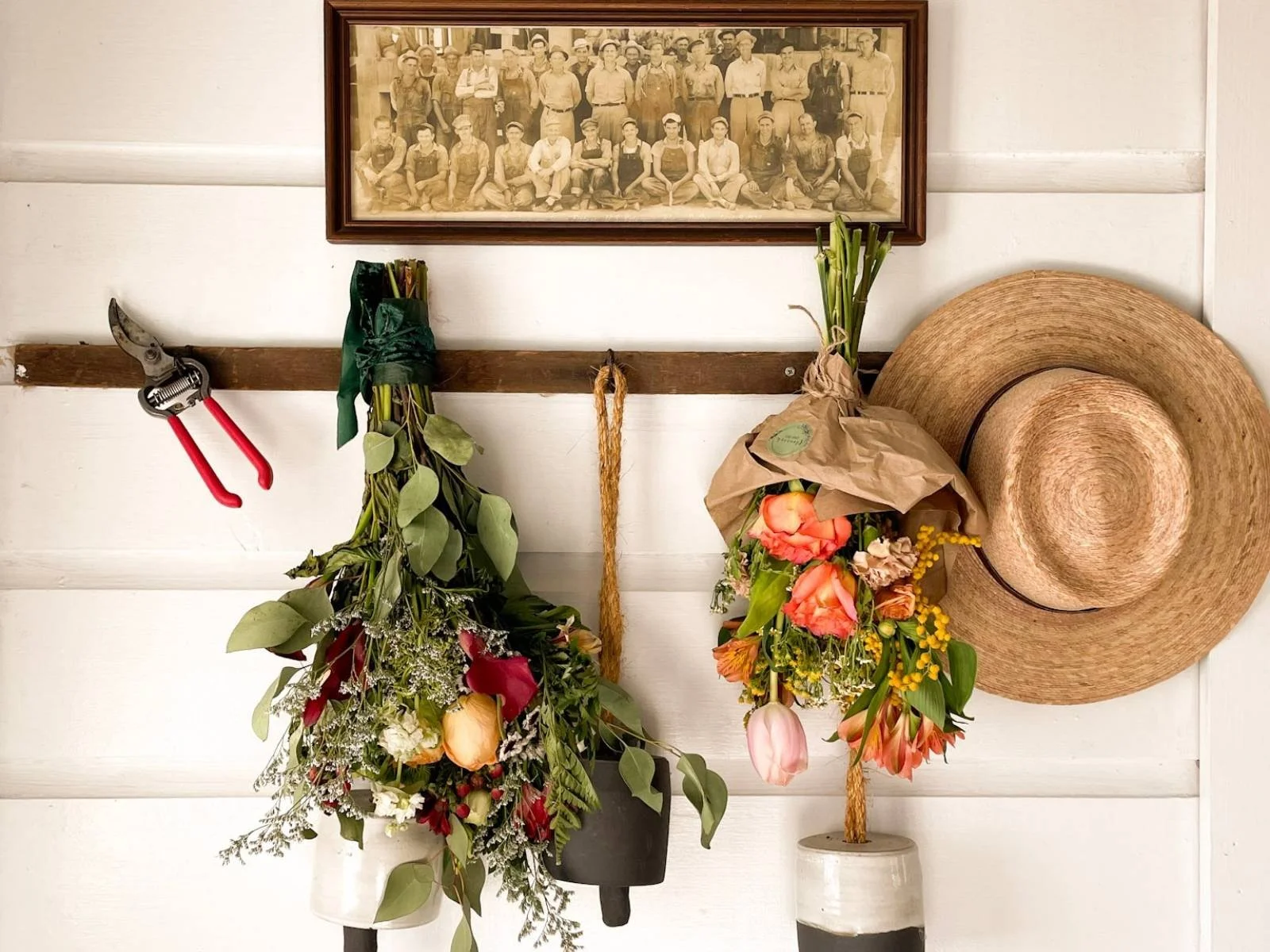Decorative display with mounted flowers, a large straw hat, a framed black-and-white family or team photo, and gardening tools on a white wall.