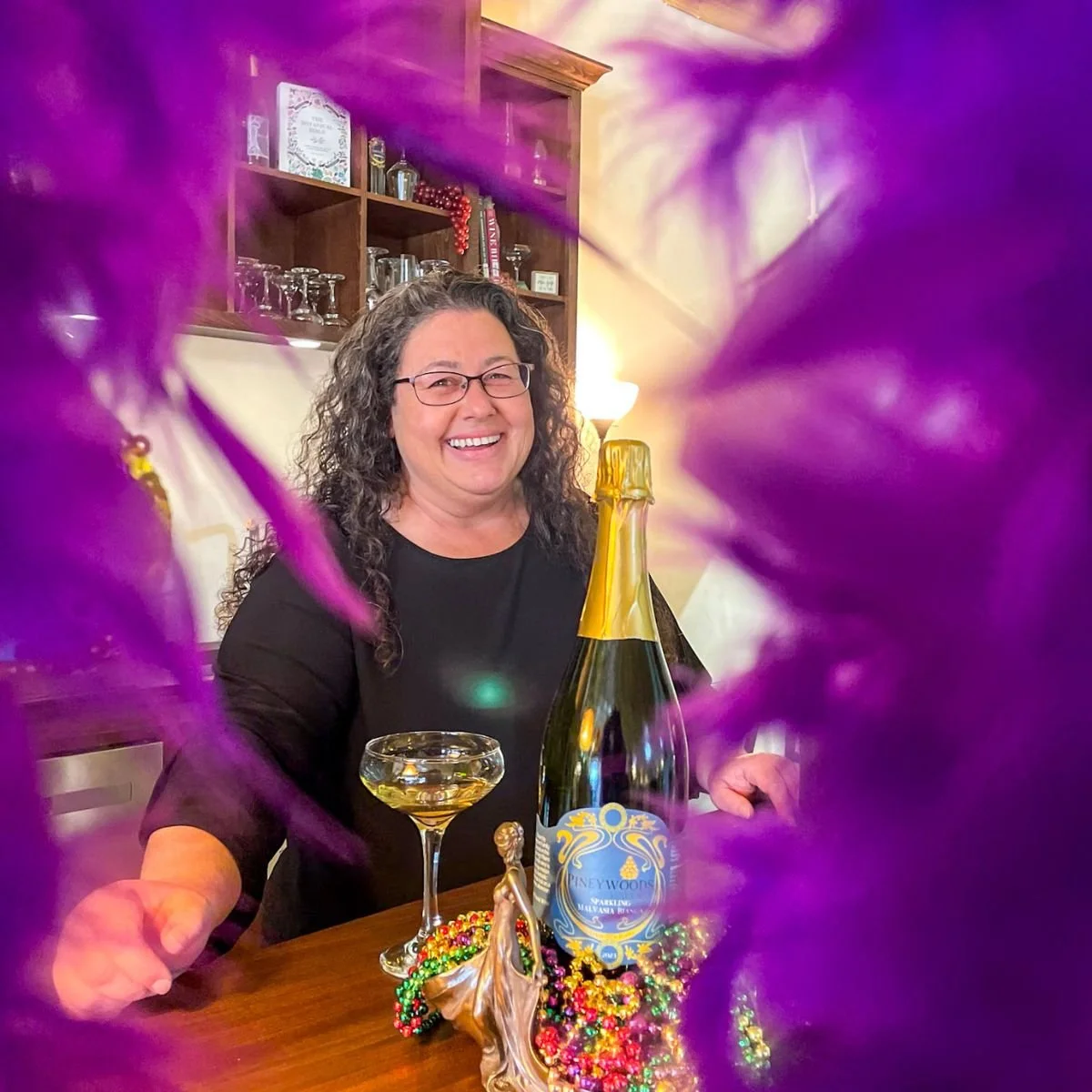 Smiling Pineywoods Vineyard staff member Karen wearing a Mardi Gras bow and holding a glass of champagne during a festive event