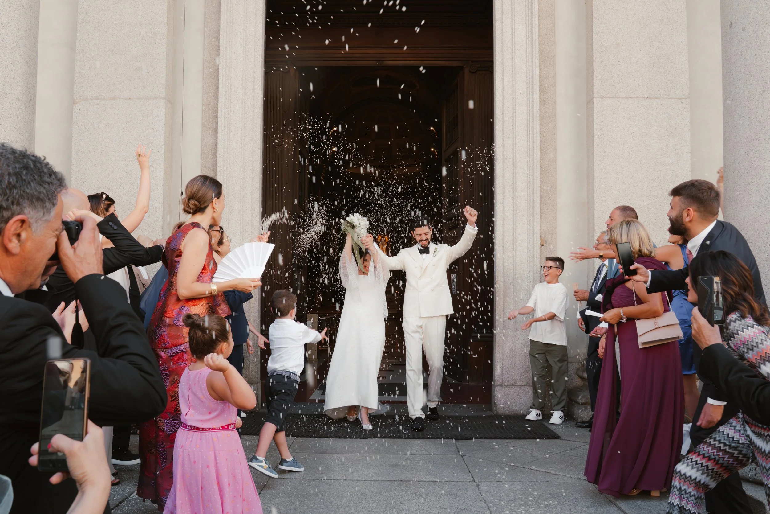 Una coppia di sposi felici mentre escono da una chiesa, circondati da amici e parenti che li festeggiano lanciando confetti.