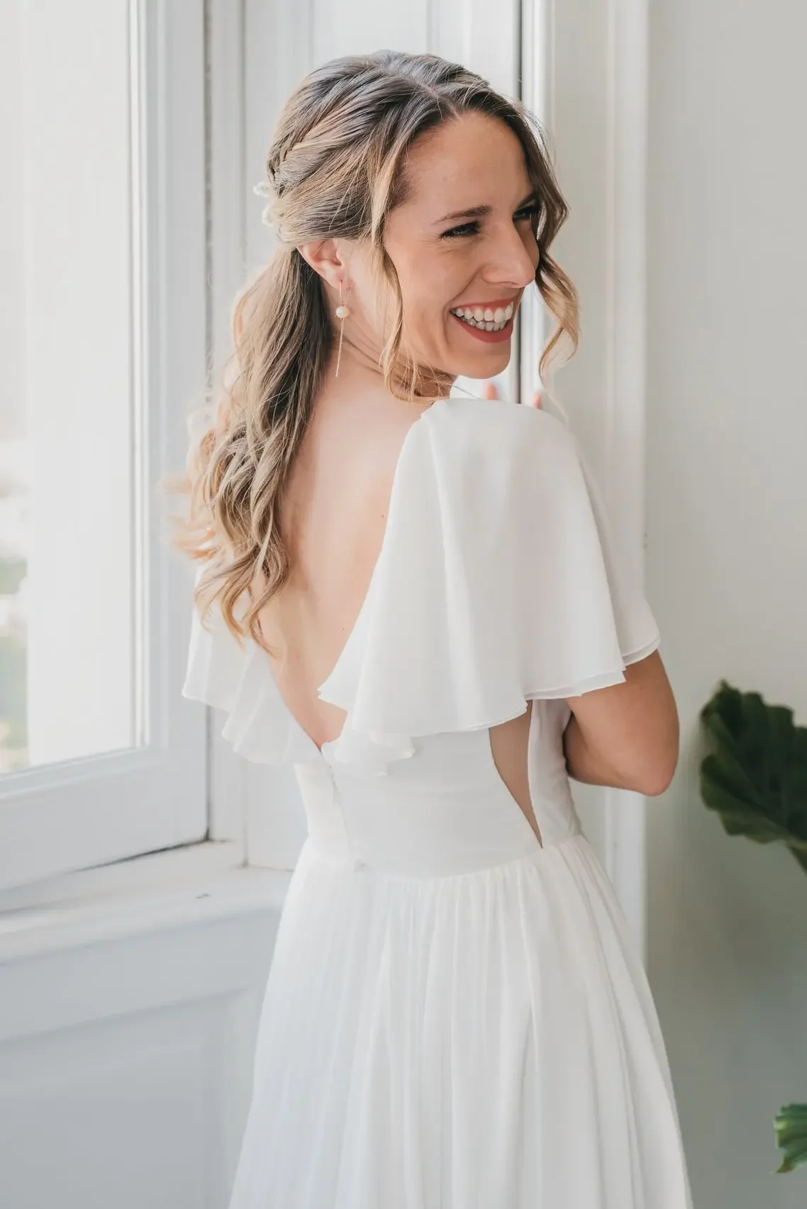 Una donna sorride, indossa un vestito bianco con maniche a ruota, in piedi vicino a una finestra con luce naturale.