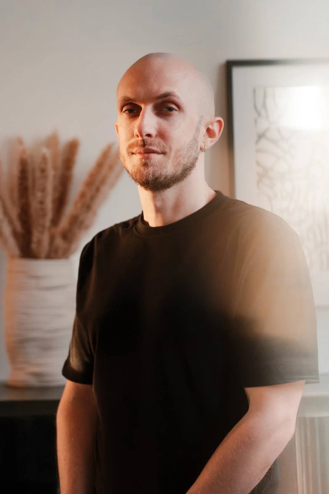 Ragazzo con pelle liscia e capelli rasati, indossa una maglietta nera, sfondo con decorazione di pampas e quadro astratto.