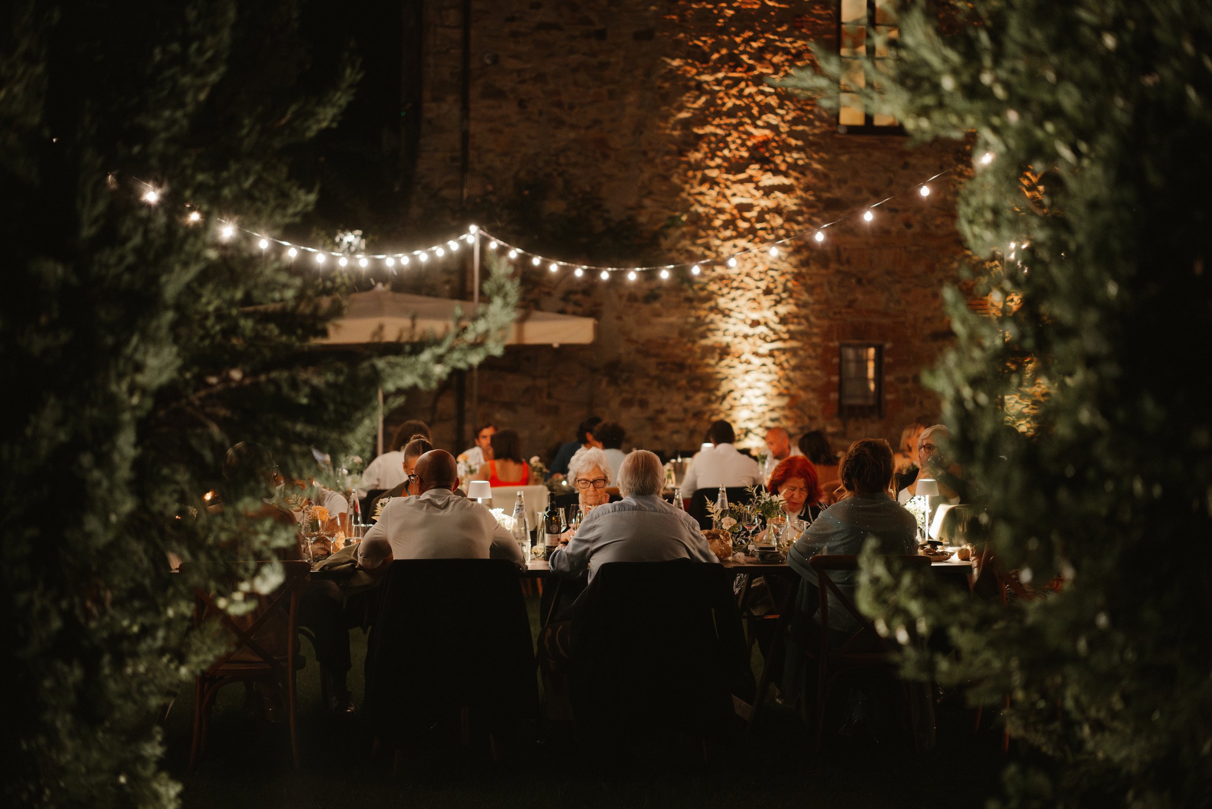 Persone sedute a un tavolo per una cena all'aperto di sera, illuminate da luci a stringa, circondate da alberi, con uno sfondo di un muro di pietra.