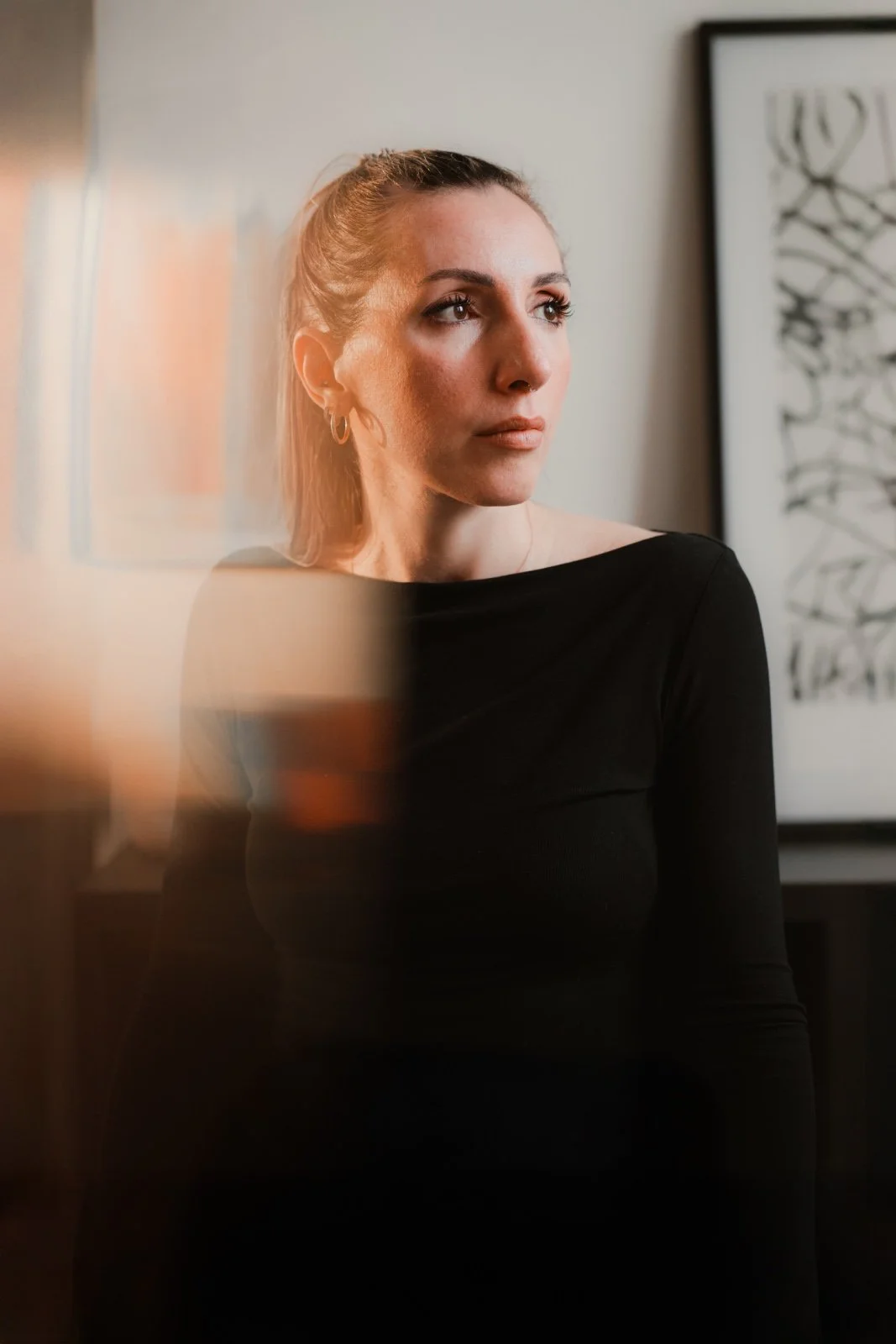 Una donna con capelli castano chiaro raccolti, indossa una maglietta nera a maniche lunghe, guarda a destra, sfondo con quadro astratto bianco e nero, luce naturale.