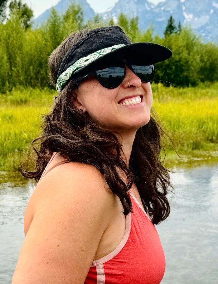 A woman smiling outdoors near a river, wearing sunglasses and a sun hat, with trees and mountains in the background.