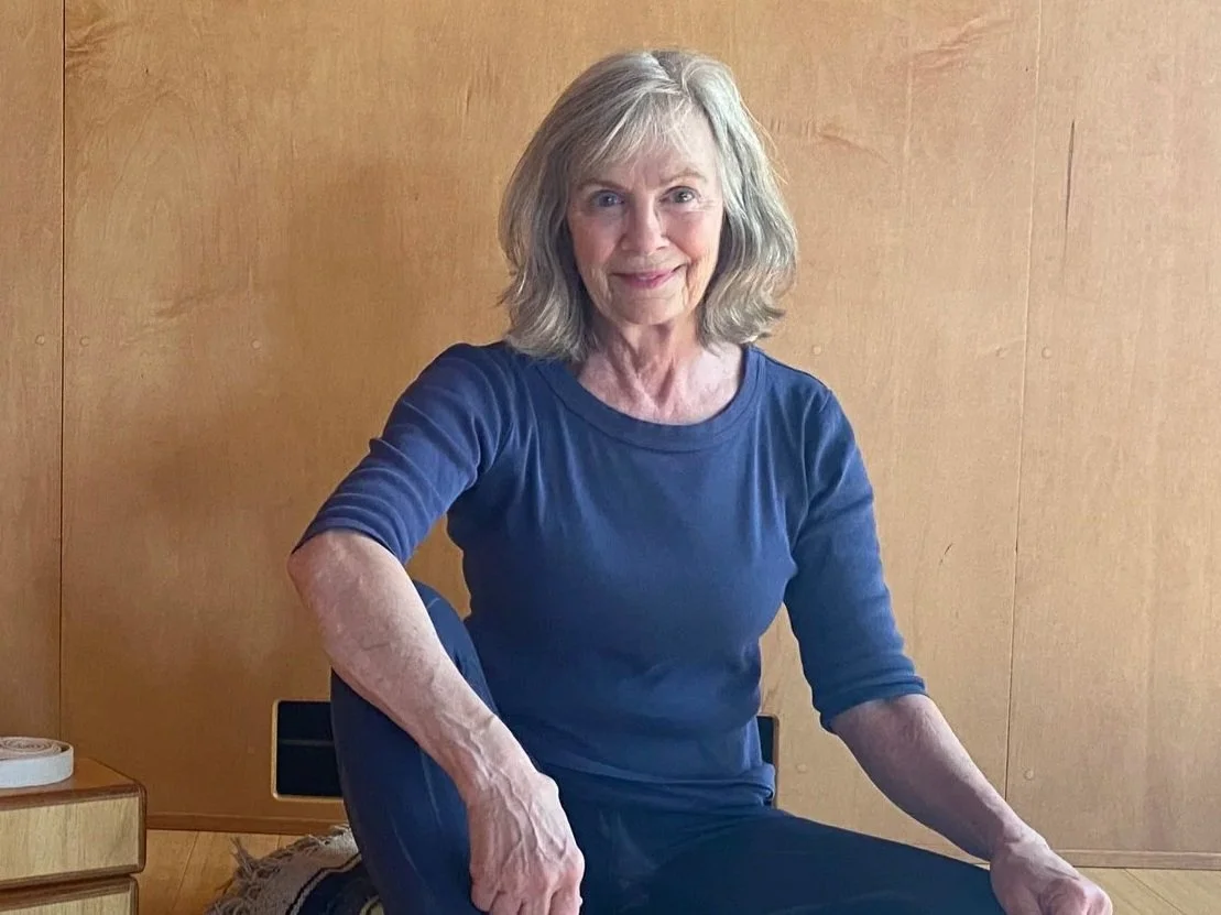 An elderly woman with gray hair and a blue long-sleeve shirt sitting on the floor against a wooden wall, smiling at the camera.