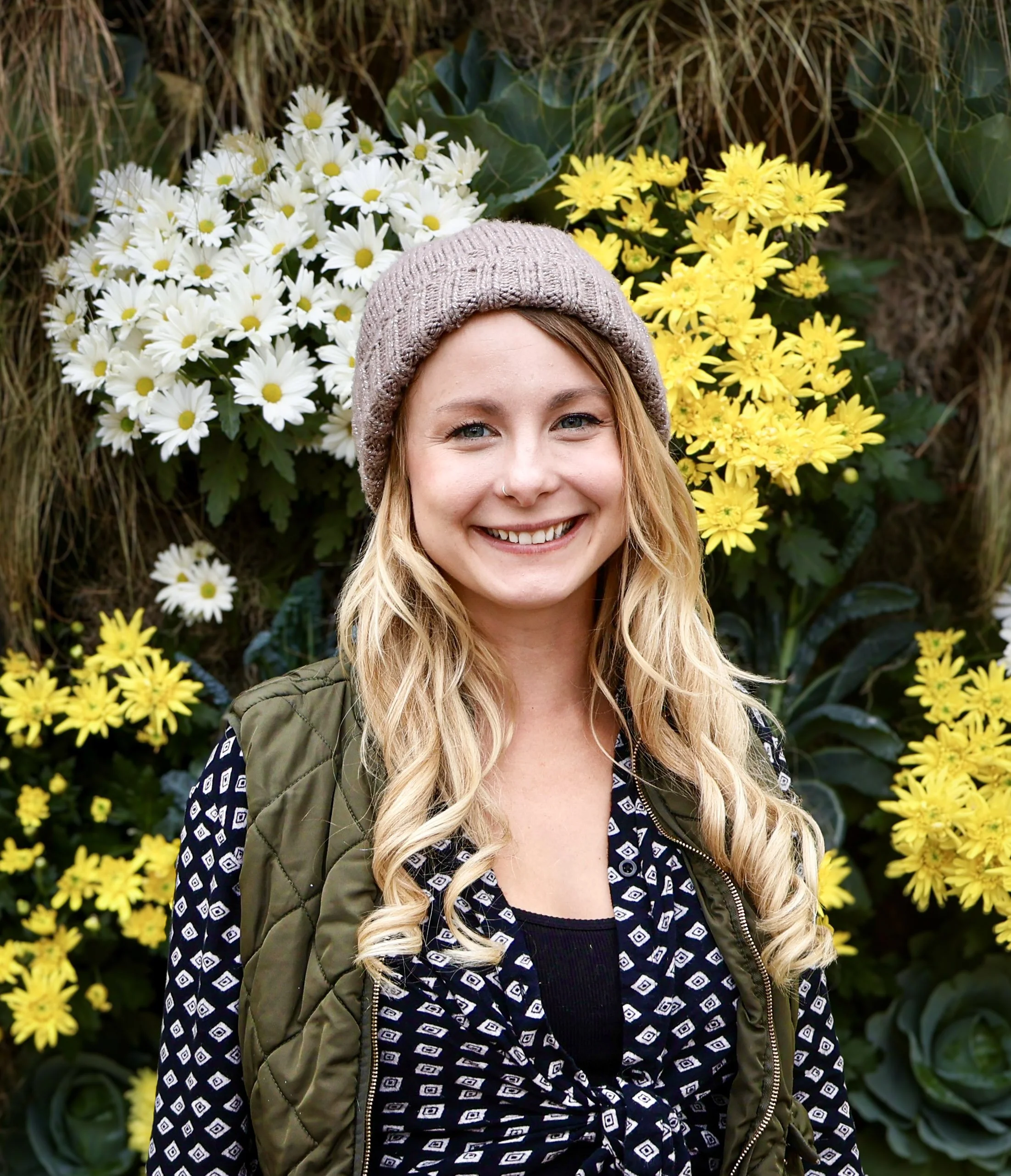 A young woman with blonde hair, blue eyes, and a nose ring, smiling, wearing a beige knit beanie, a patterned black and white top, and a green quilted jacket, in front of white and yellow flowers.