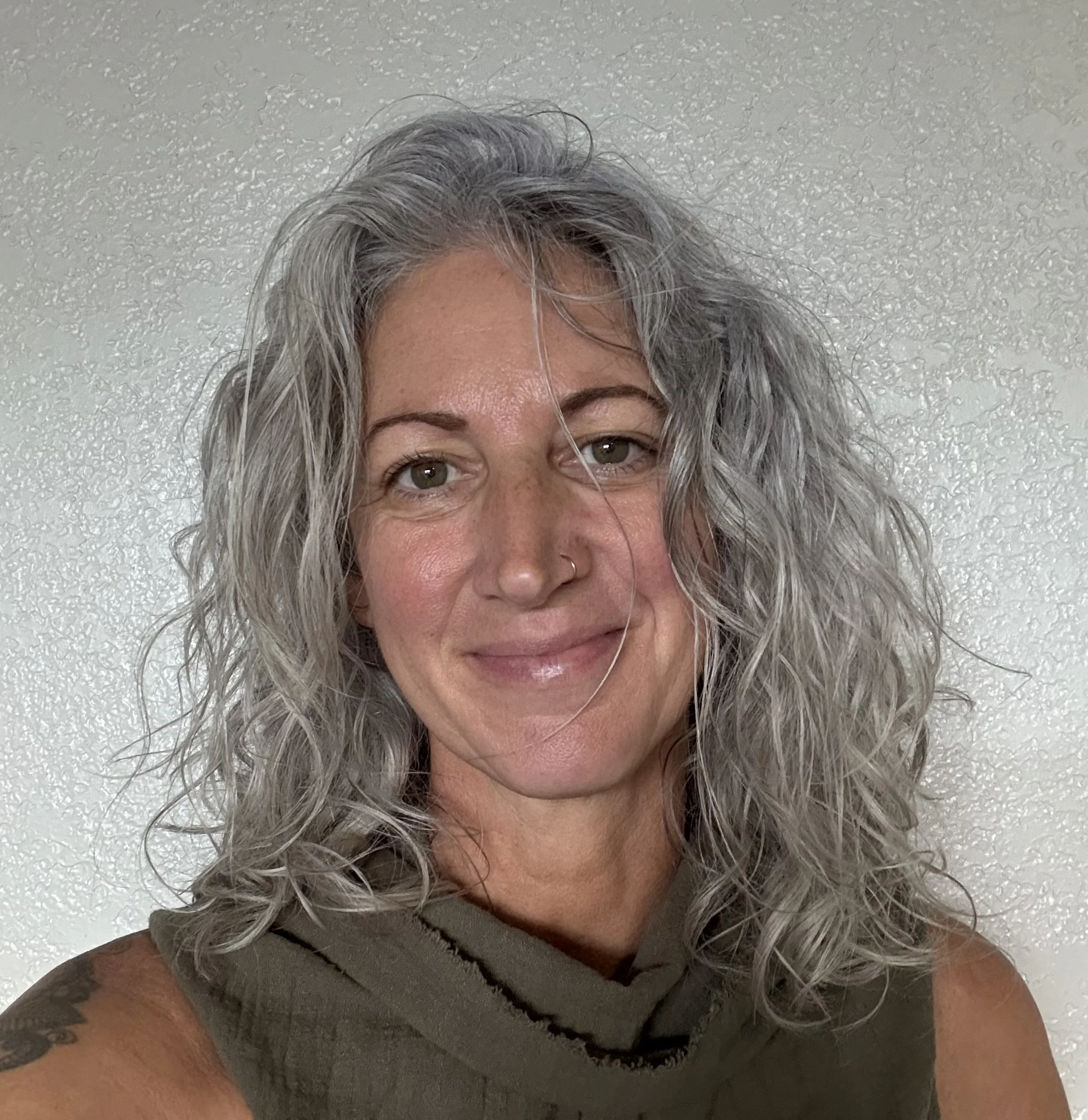 A woman with curly gray hair smiling at the camera, wearing a sleeveless olive green top, standing against a textured white wall.