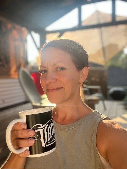 A woman with short brown hair holding a black mug with white text, smiling outdoors with sunlight and patio furniture in the background.
