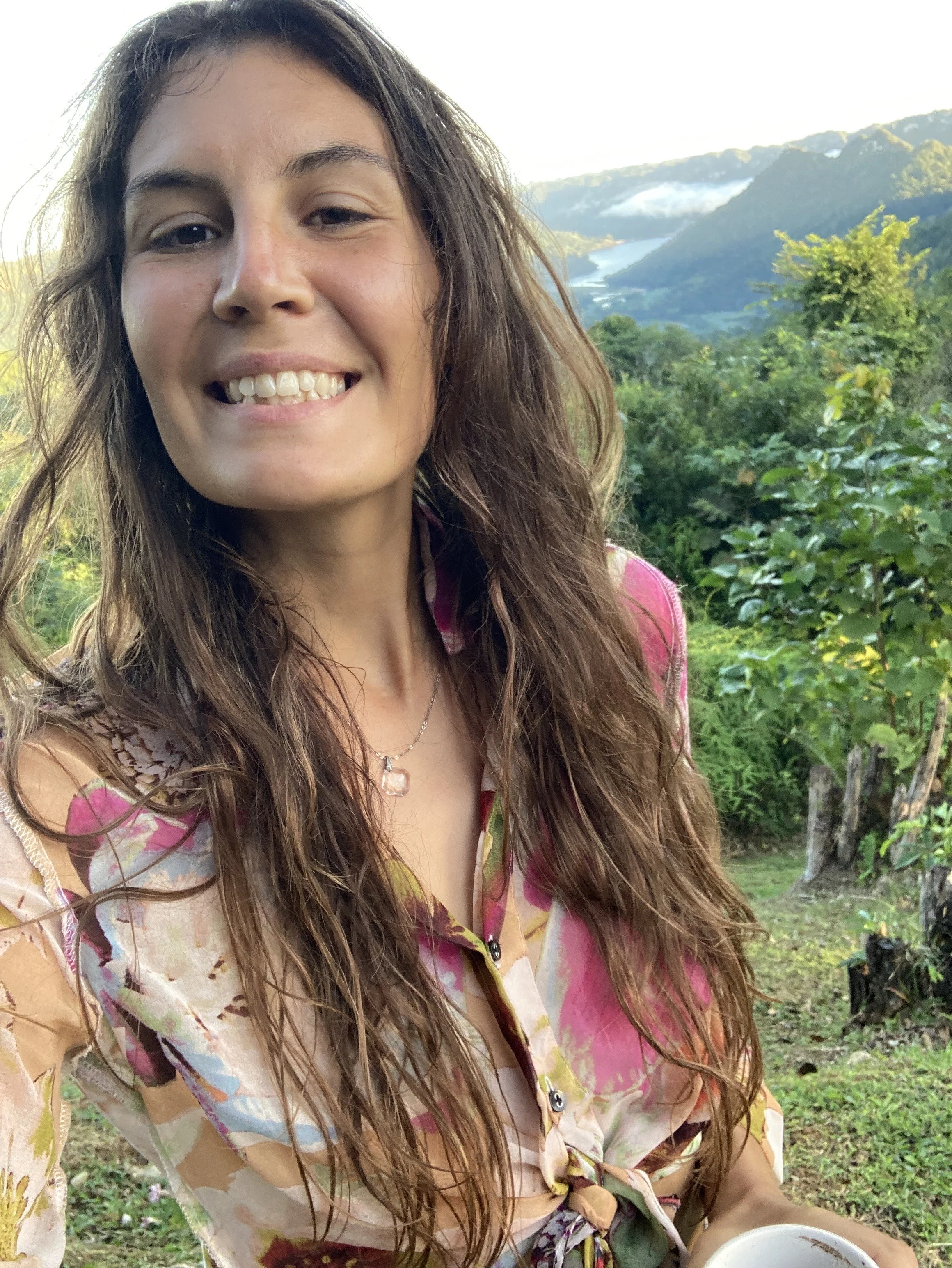 A smiling woman with long, wavy hair outdoors, holding a cup, with a scenic background of green trees, hills, and a distant river or lake under a partly cloudy sky.