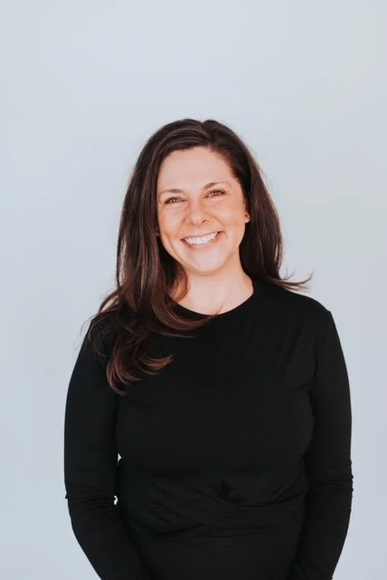 Smiling woman with long brown hair wearing a black top against a light background.