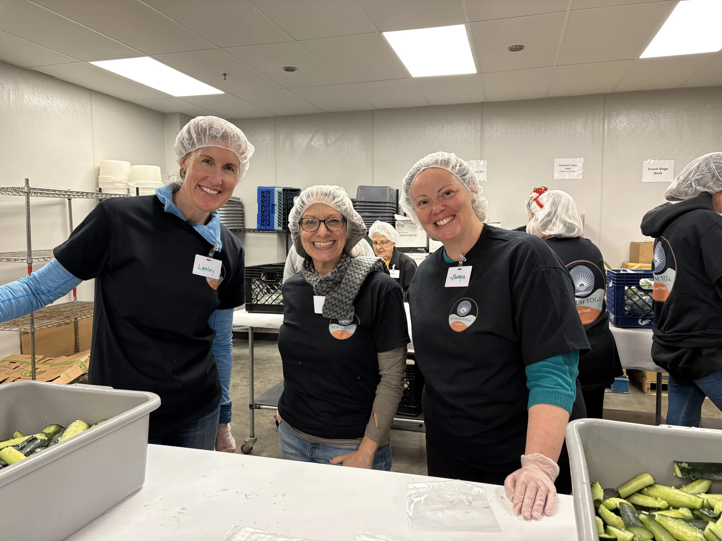 Three women in hairnets and black shirts with name tags smiling at the camera in a food packaging or packing facility.