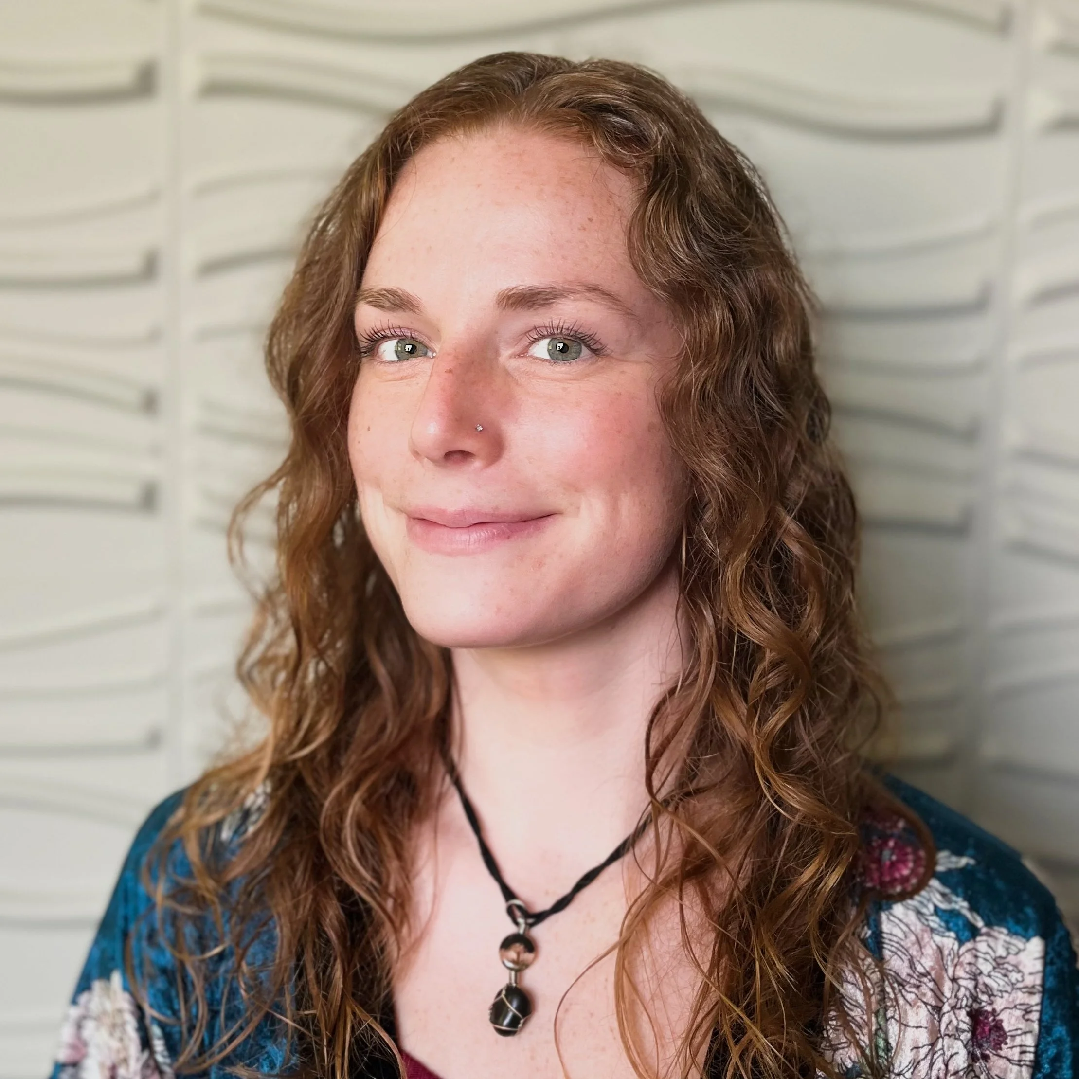 A woman with long, curly red hair, light green eyes, and a nose piercing, smiling softly, wearing a black necklace with a pendant, standing against a textured white background.
