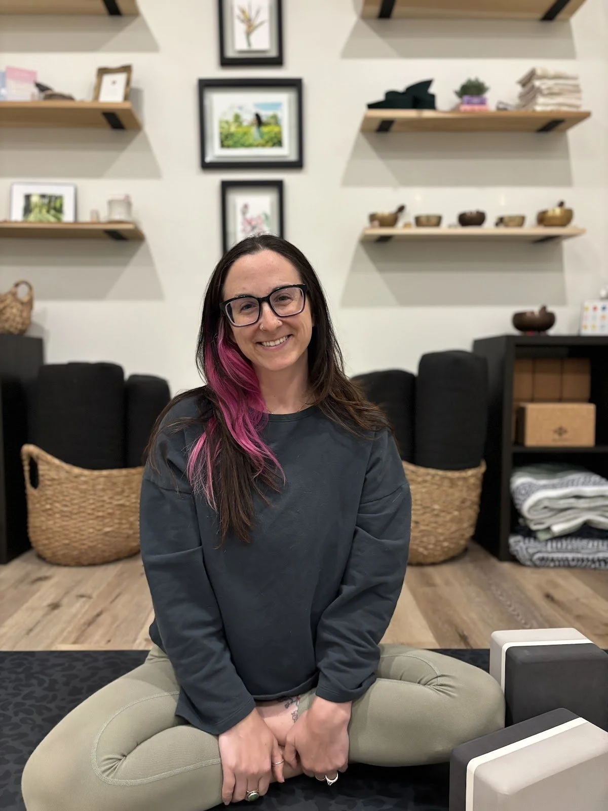 A woman with long brown hair with pink highlights, wearing glasses and a dark long-sleeve shirt, sitting cross-legged on a black cushion in a cozy, well-decorated room with artwork and shelves in the background.
