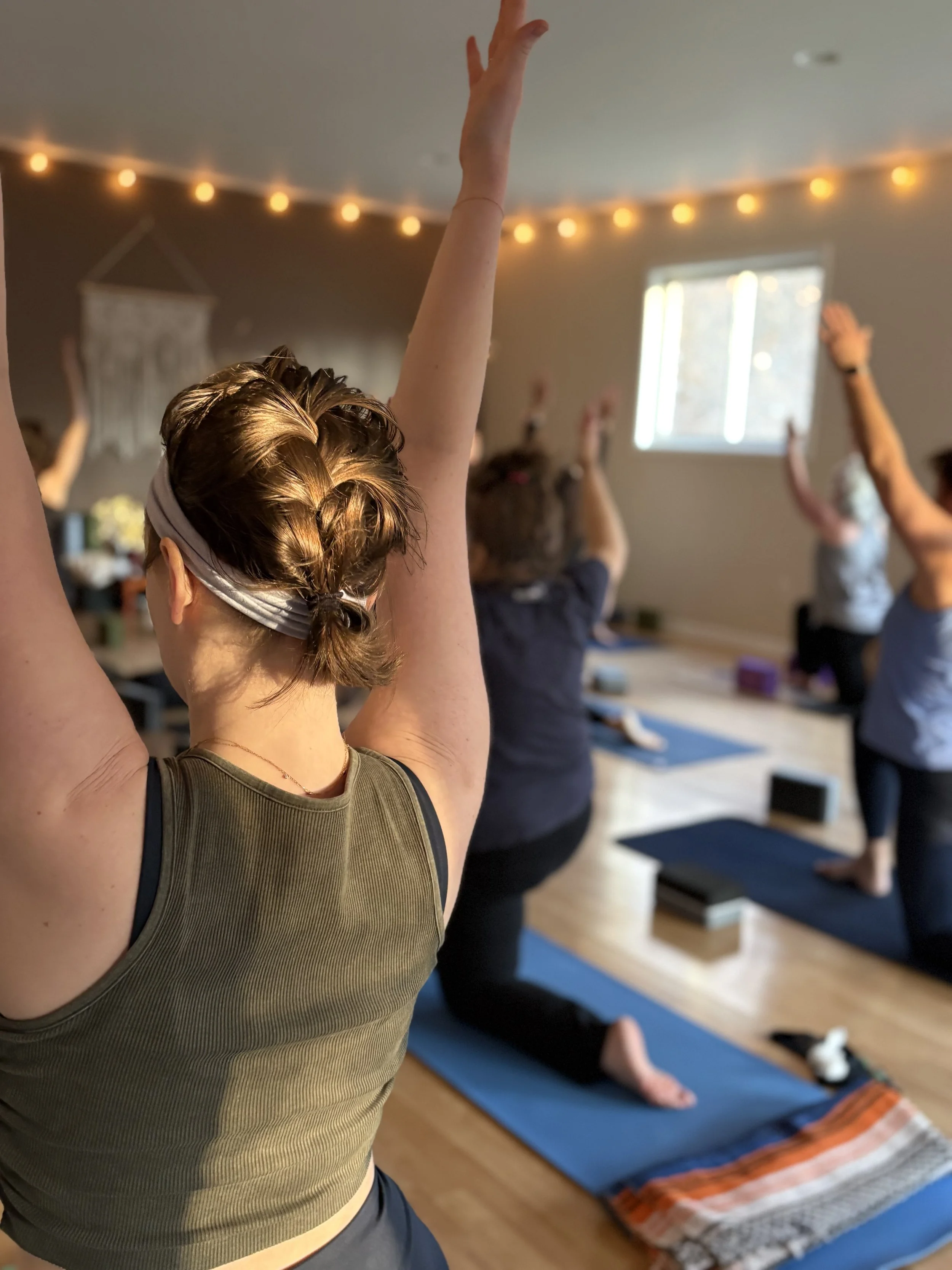 A group of people practicing yoga in a cozy indoor studio with string lights and a window, with participants in various yoga poses.
