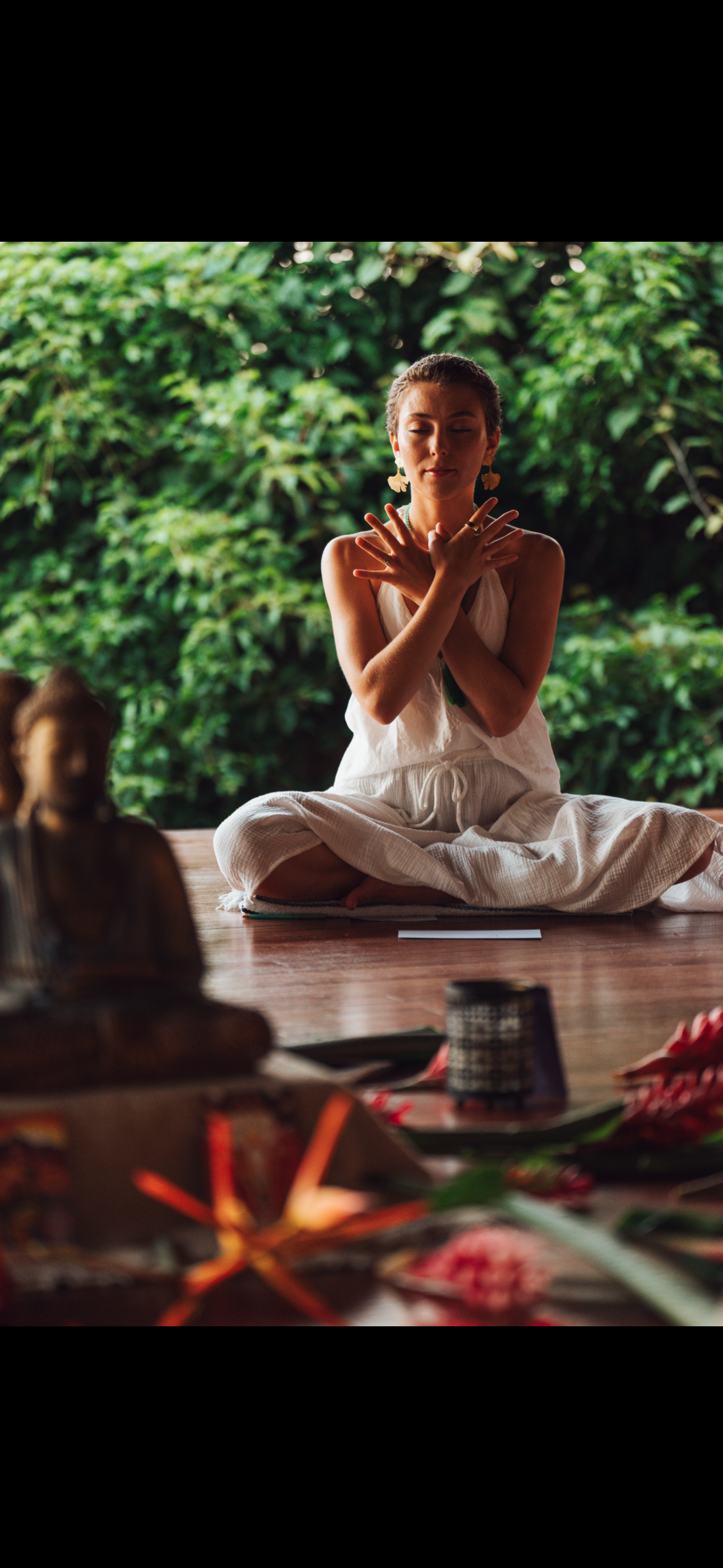 A woman practicing yoga outdoors seated cross-legged on a wooden deck with lush green foliage in the background. She is wearing a white sleeveless dress and gold earrings, with her hands crossed over her chest and eyes closed, in a peaceful pose. Various objects and a Buddha statue are visible in the foreground.