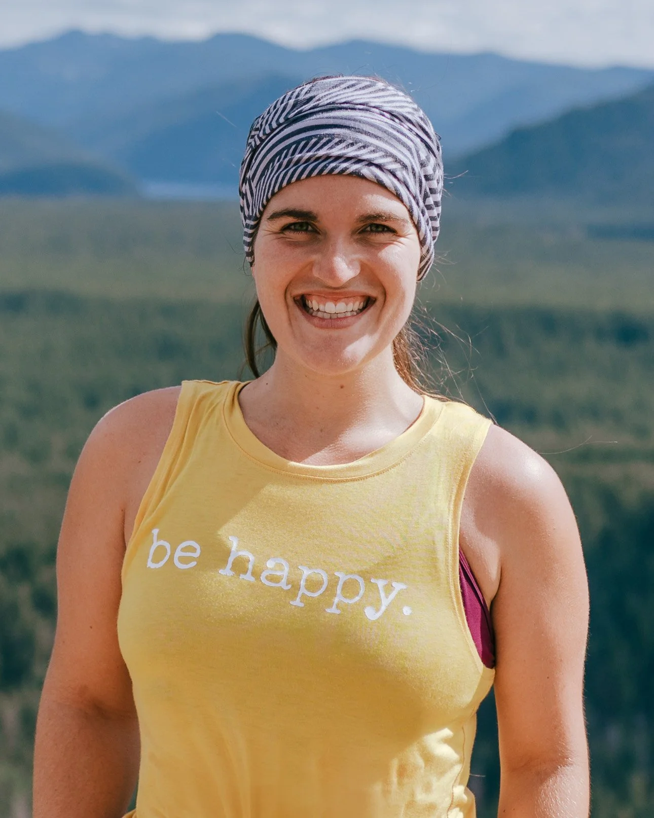 Smiling woman wearing a yellow tank top with the words 'be happy.' and a striped headscarf, outdoors with mountains and a blue sky in the background.