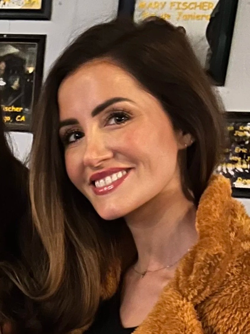 A woman with long brown hair, smiling, wearing makeup, a black top, and a tan furry jacket, in front of a wall with framed posters.