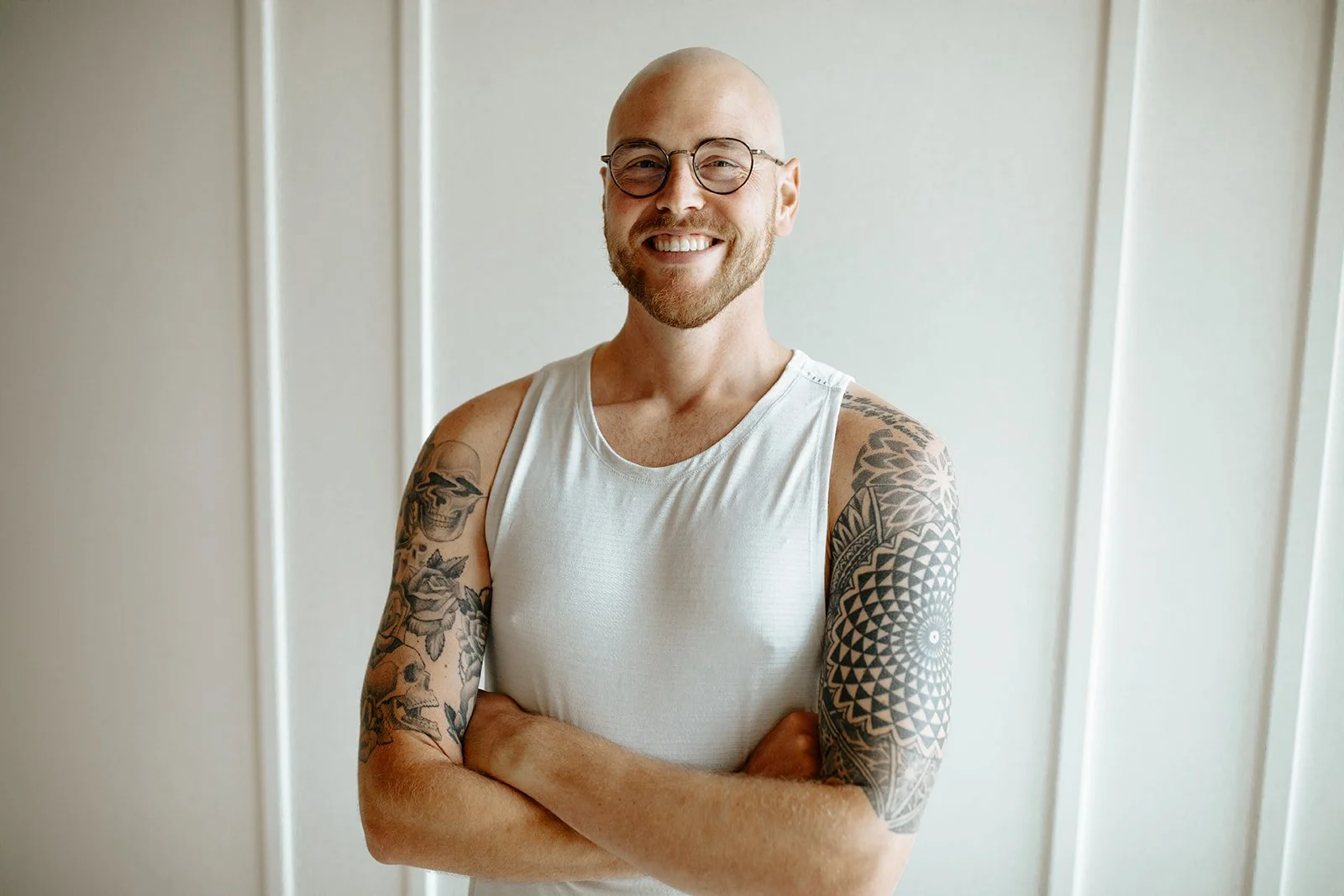 A smiling bald man with glasses wearing a sleeveless white shirt, crossed arms, showcasing tattoo sleeves on both arms, standing in front of a light-colored wall.