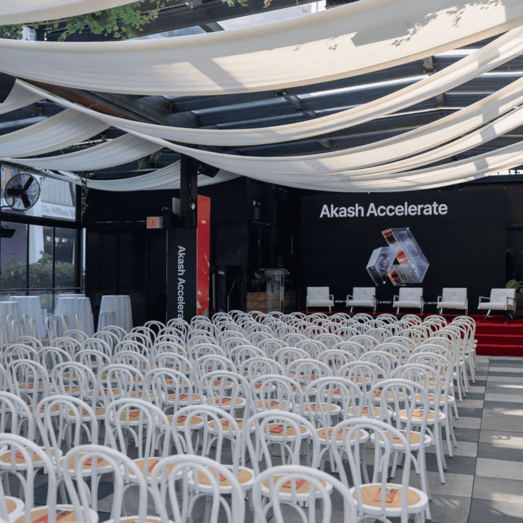 Empty chairs arranged in rows in an indoor venue with a stage that has a black backdrop displaying the words "Akash Accelerate" and a hexagonal graphic. White drapery decorates the ceiling, and there is a large window on the left side.