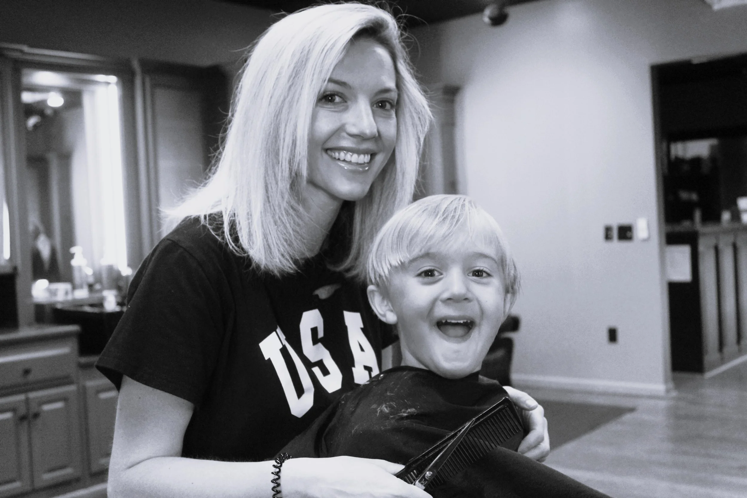 A black and white photo of a woman with shoulder-length blonde hair smiling and a young boy with blonde hair smiling in a room with cabinets and a stairs in the background.