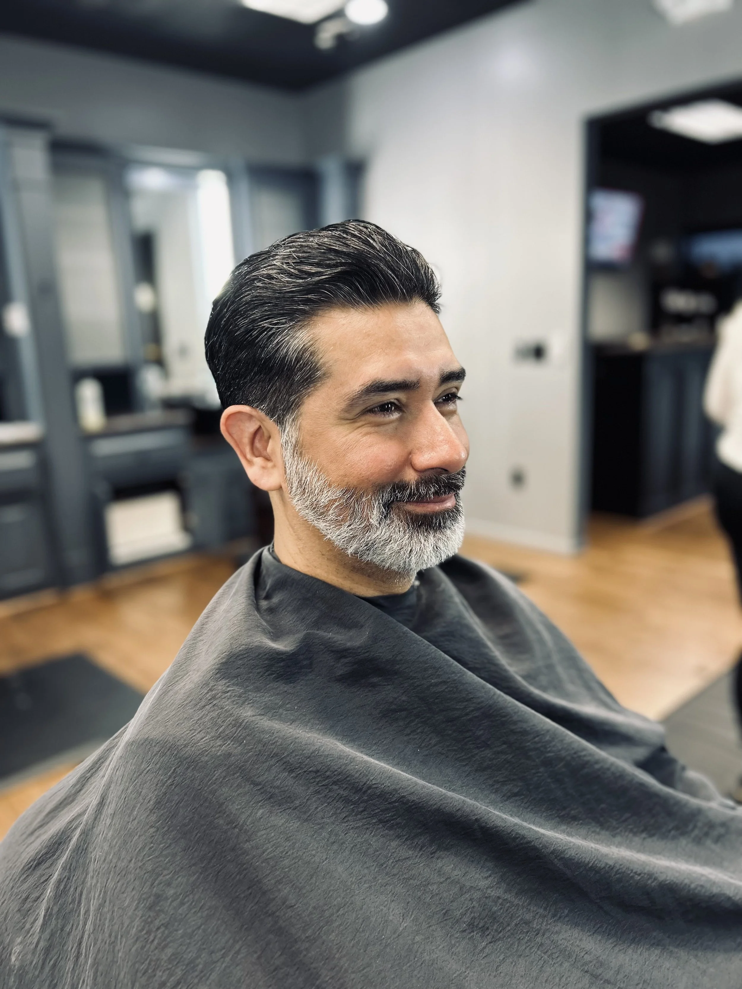 A smiling man with a groomed beard and dark, styled hair sitting in a barbershop chair wearing a black cape.