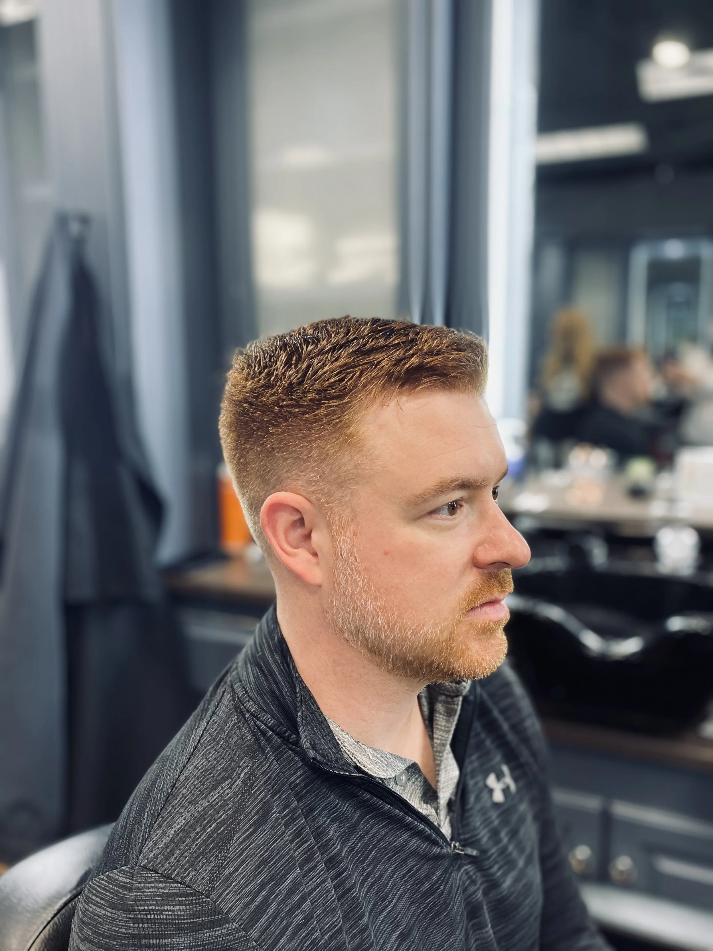 Man with short, styled brown hair and a beard sitting in a barbershop chair, wearing a black Under Armour jacket.