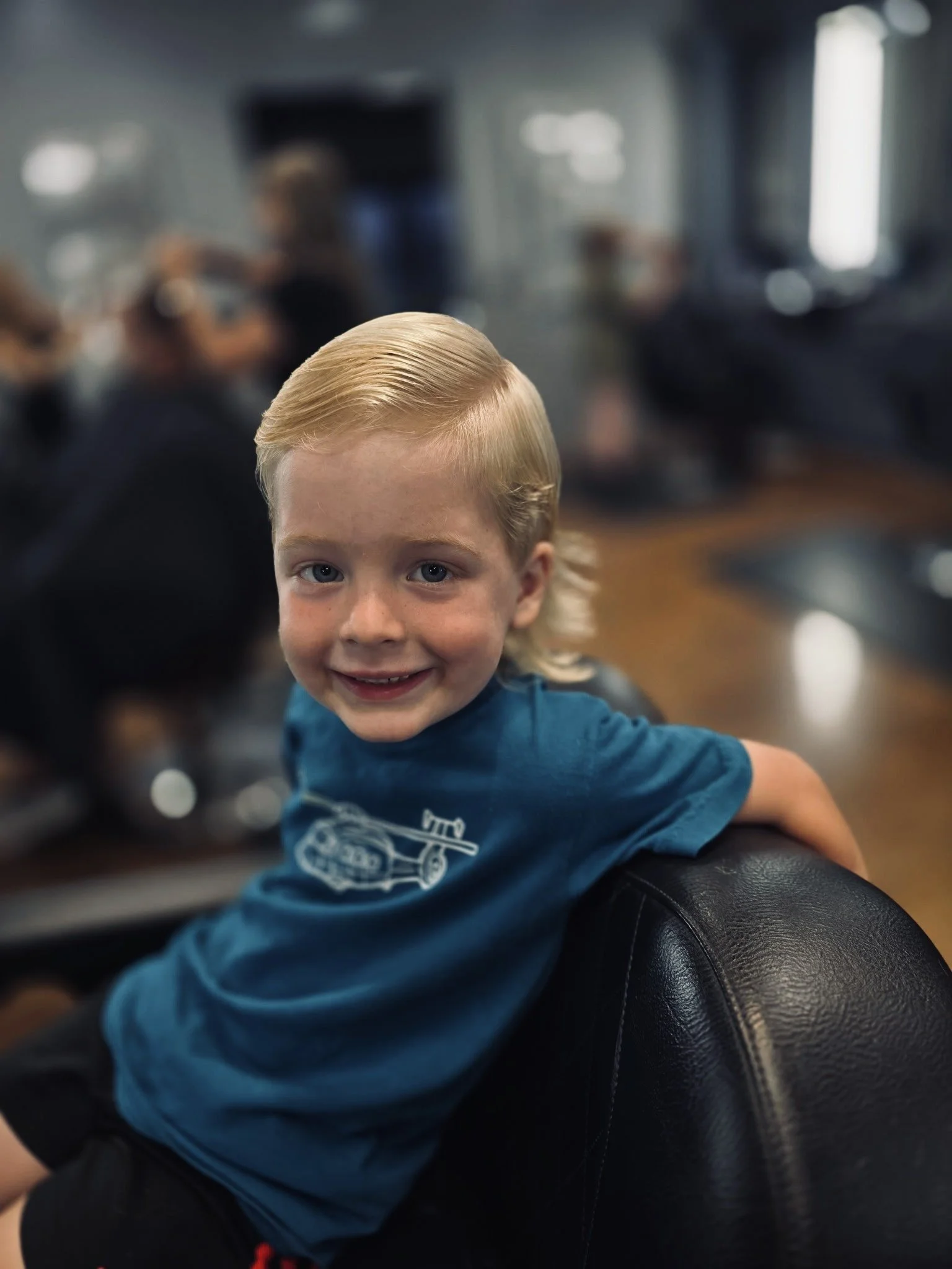 A young child with blonde hair smiling at the camera, sitting on the back of a black leather chair, with a blurred room and people in the background.
