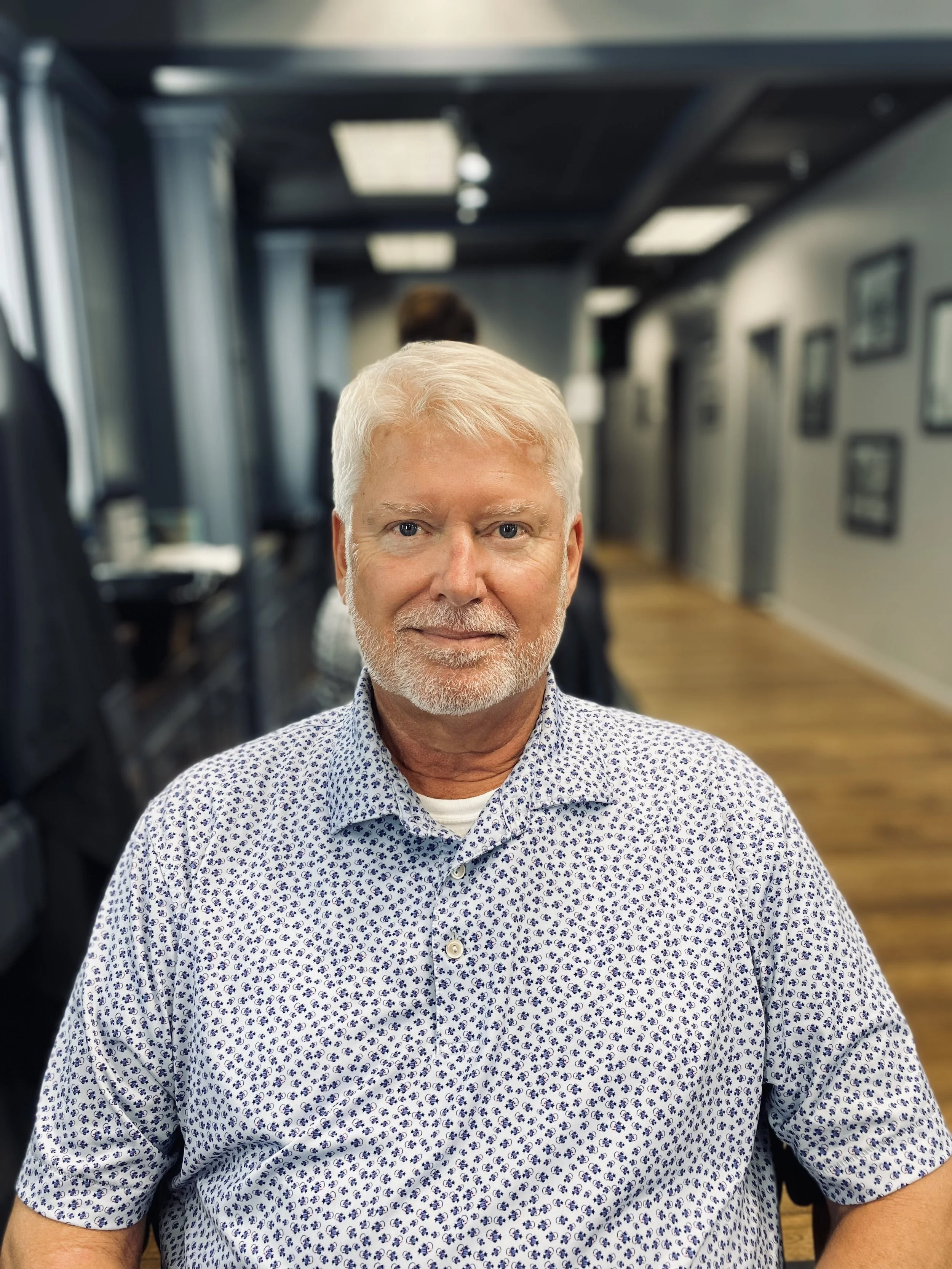 A mature man with white hair and beard wearing a patterned button-up shirt, sitting in an indoor office or lounge area with framed pictures on the wall behind him.