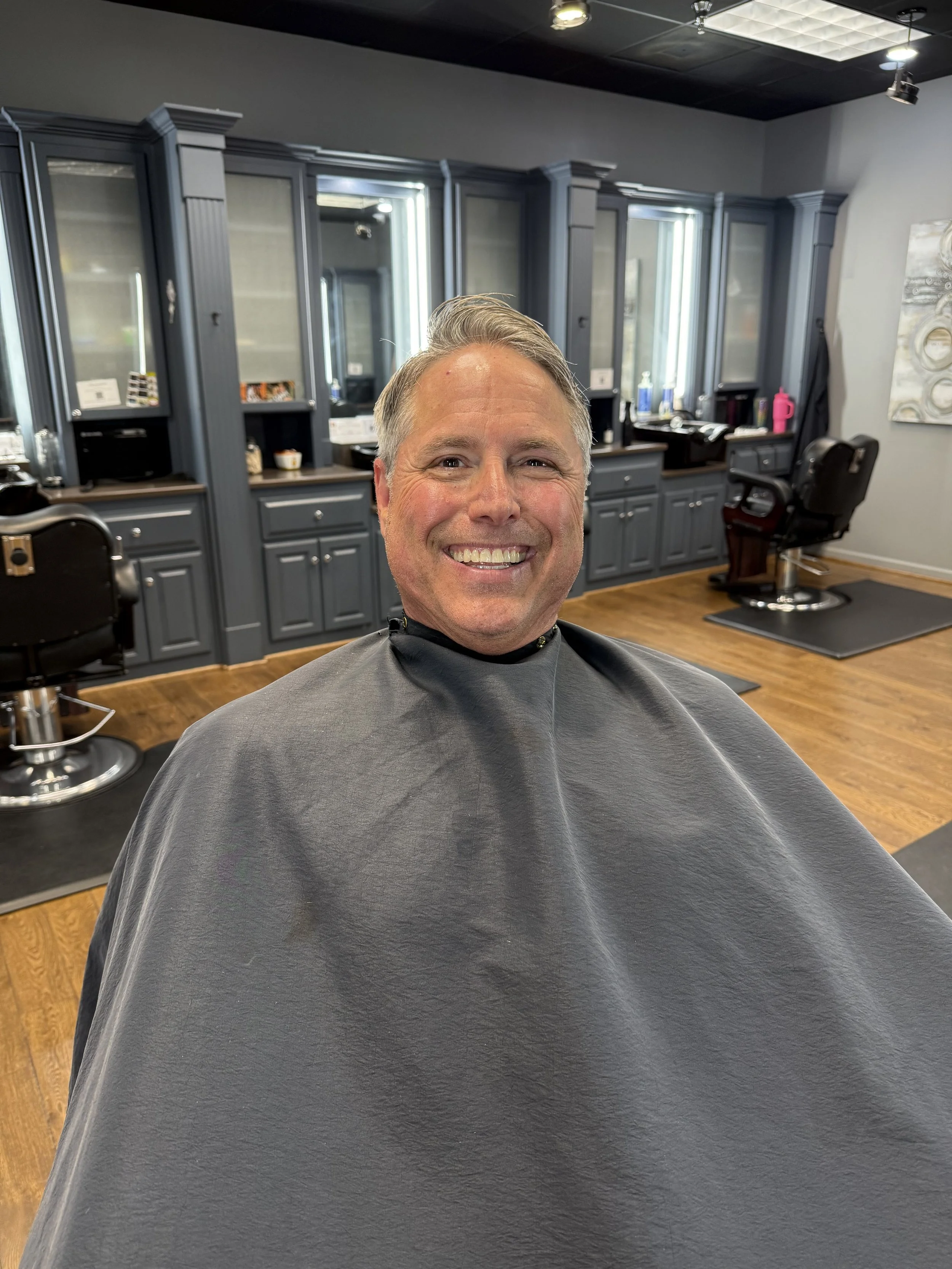 A man smiling in a barbershop, wearing a black barber cape, with dark blue cabinets and mirrors in the background.