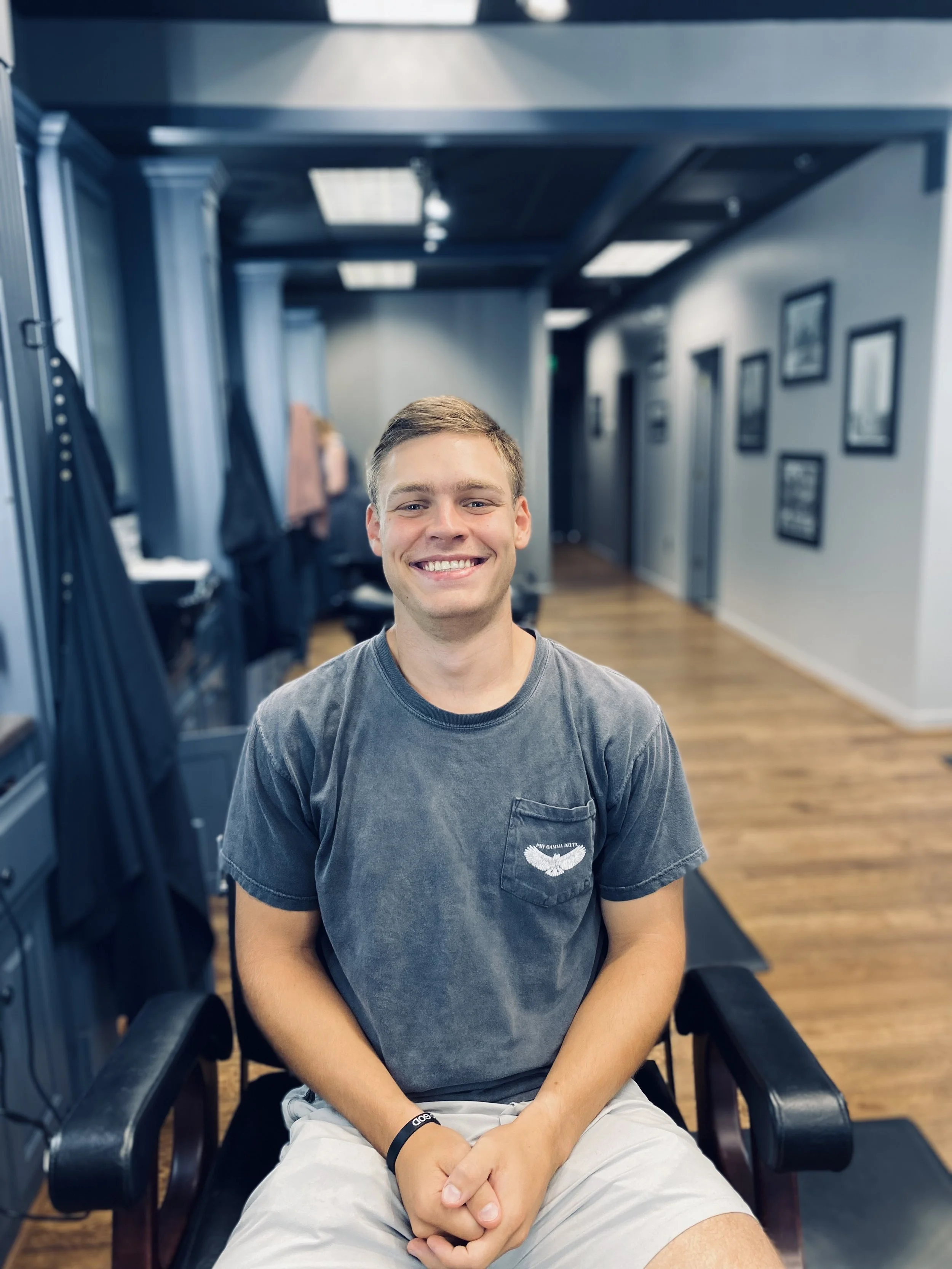A smiling young man sitting in a barber's chair inside a hair salon with framed pictures on the wall in the background.