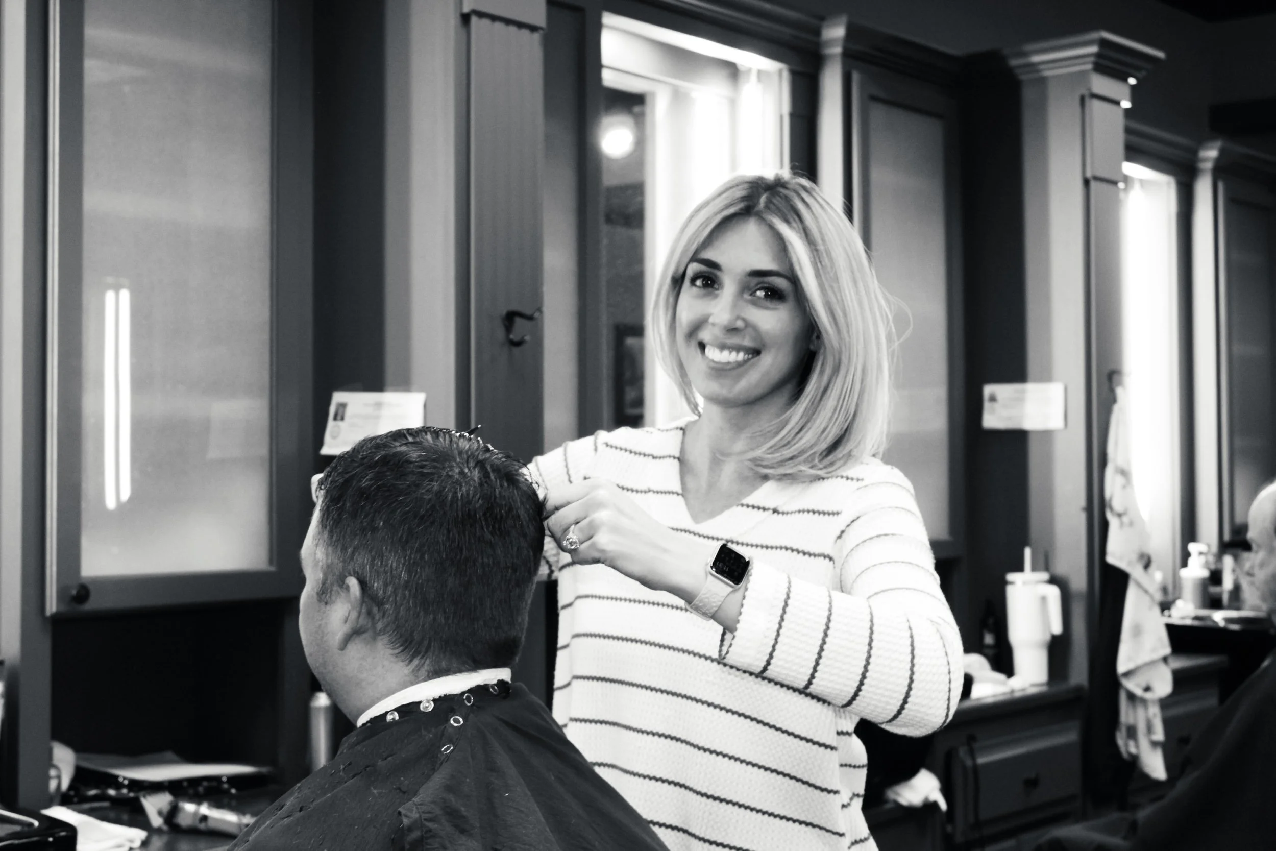 A woman with shoulder-length blonde hair smiling while cutting a man's hair at a barbershop.