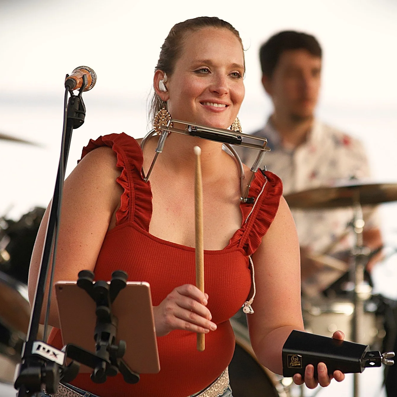A woman smiling, wearing a red dress with ruffled straps, playing a percussion instrument, with a man in the background playing the drums.