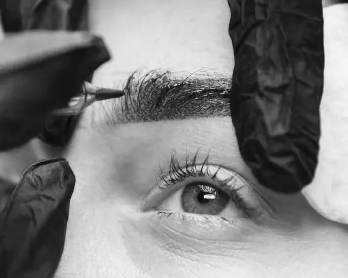 Close-up of a person receiving an eyebrow tattooing or microblading procedure, with a technician wearing black gloves applying pigment to the eyebrow area.