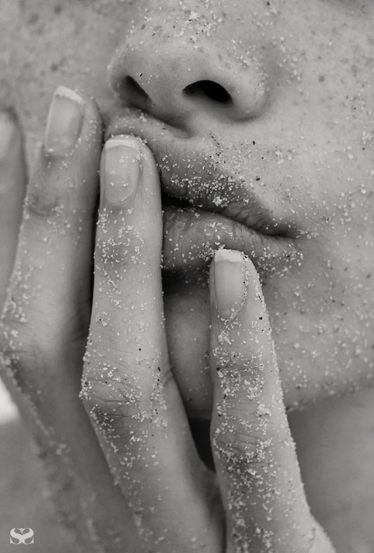A close-up of a person's face covered in sand, with fingers near the lips, emphasizing the lips and nose.