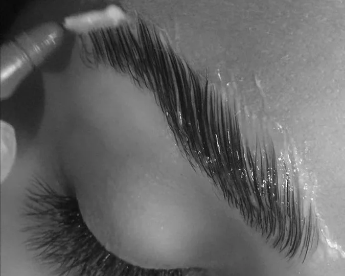Close-up of a person's closed eye with long eyelashes, and someone applying brow lamination product with a small brow tool.