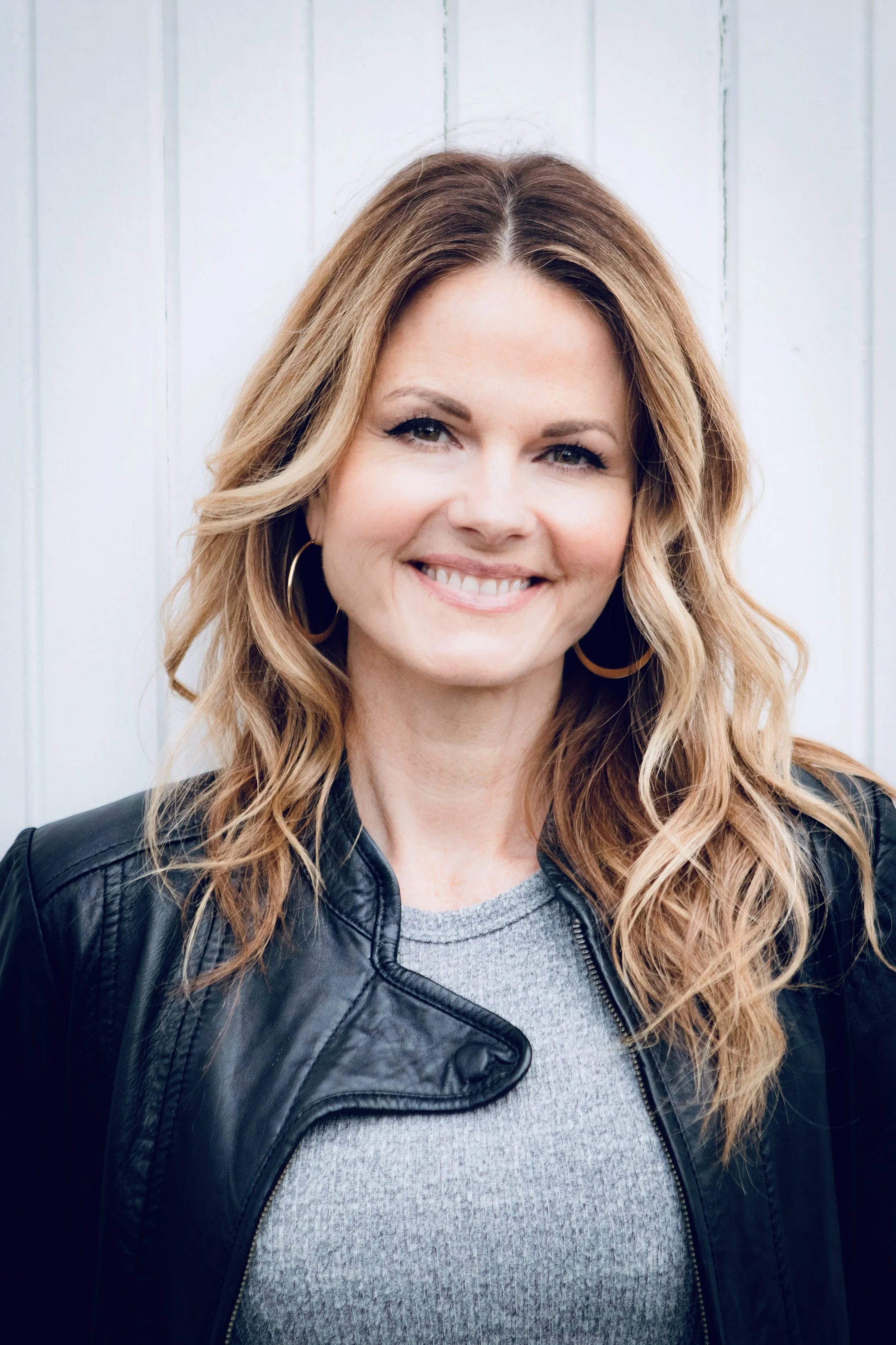 Raechelle in a leather jacket, smiling, standing in front of a shiplap wall.