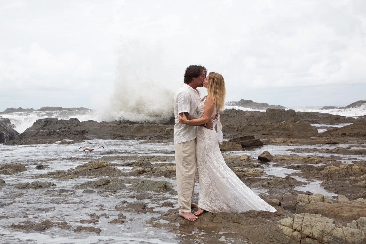 Raechelle and her new husband on thier wedding day in front of a crashing wave.  They are barefoot, in their wedding attire, kissing on a rock shelf in front of the ocean.