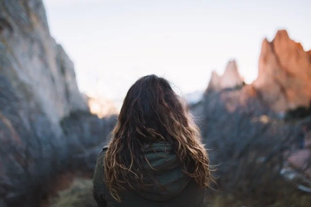 A woman sitting in the mountains looking out over a valley and mountain range in the distance.