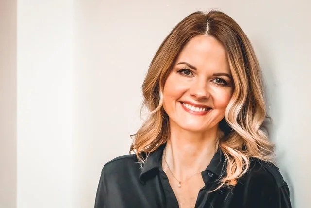 Raechelle is standing against a white wall background.  She is smiling with curled blonde hair and a button up black blouse.